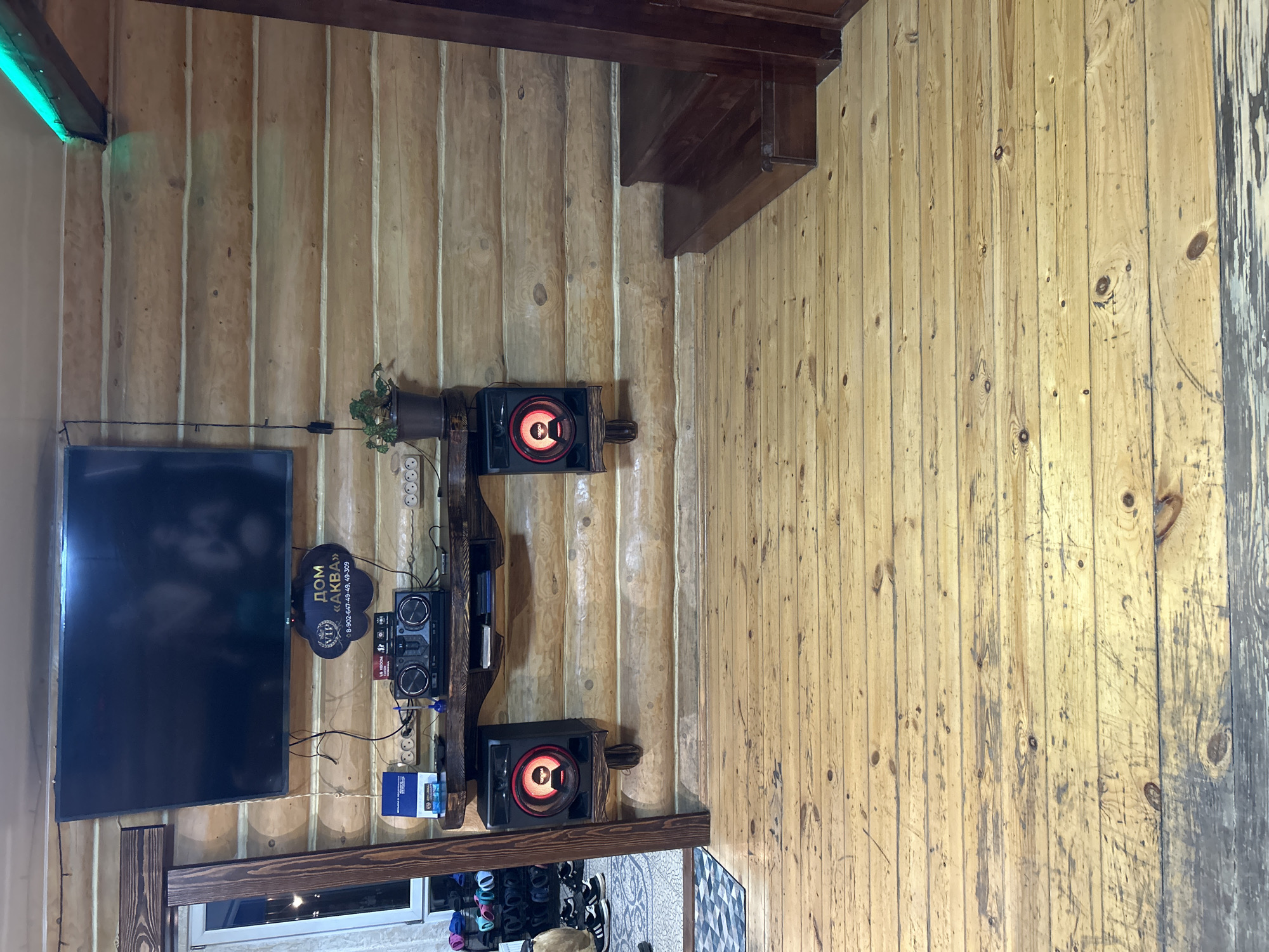The image shows an interior wall of a log cabin-style room. The wall is made of light-colored, vertically arranged logs. A flat-screen TV is mounted on the wall, along with a shelf unit holding a stereo system, speakers, a plant, and a sign in Russian that likely indicates the location as Chaikovsky, Russia ("Дом Аква"). Below the TV, there's a wooden beam-like structure with a window below it. The floor is partially visible and shows footwear on a floor with a patterned rug. Stairs are visible at the top right of the image. The scene is well-lit, though the lighting appears to vary across the wall.