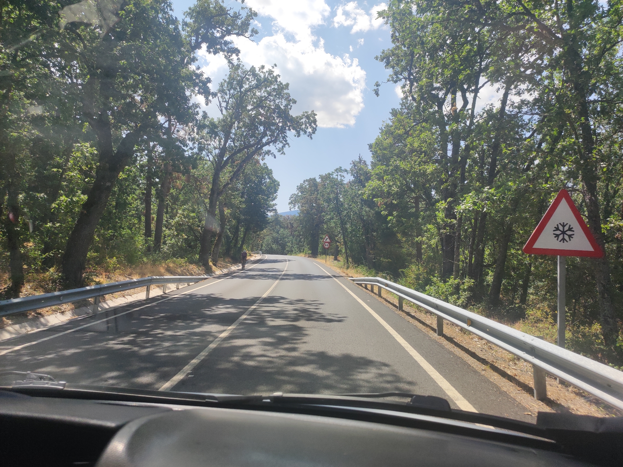 The image captures a view from inside a vehicle driving on a paved road through a forest. The road is asphalt and has white lane markings. On the right side of the road, there is a metal guardrail and a triangular road sign with a red border and a snowflake symbol, indicating icy conditions. Further down the road, another circular sign shows "50". Trees line both sides of the road, with dense foliage and some patches of sunlight filtering through. The sky above is blue with scattered white clouds. In the distance, a person on a bicycle is visible on the left side of the road. The terrain appears to be slightly inclined, suggesting an uphill or downhill stretch of road. The overall scene depicts a scenic drive on a clear day within a wooded area, likely in a rural or mountainous region. Based on the context of the prompt, this road is situated in the vicinity of Real Sitio de San Ildefonso, Spain, a location known for its royal palace and extensive forests.