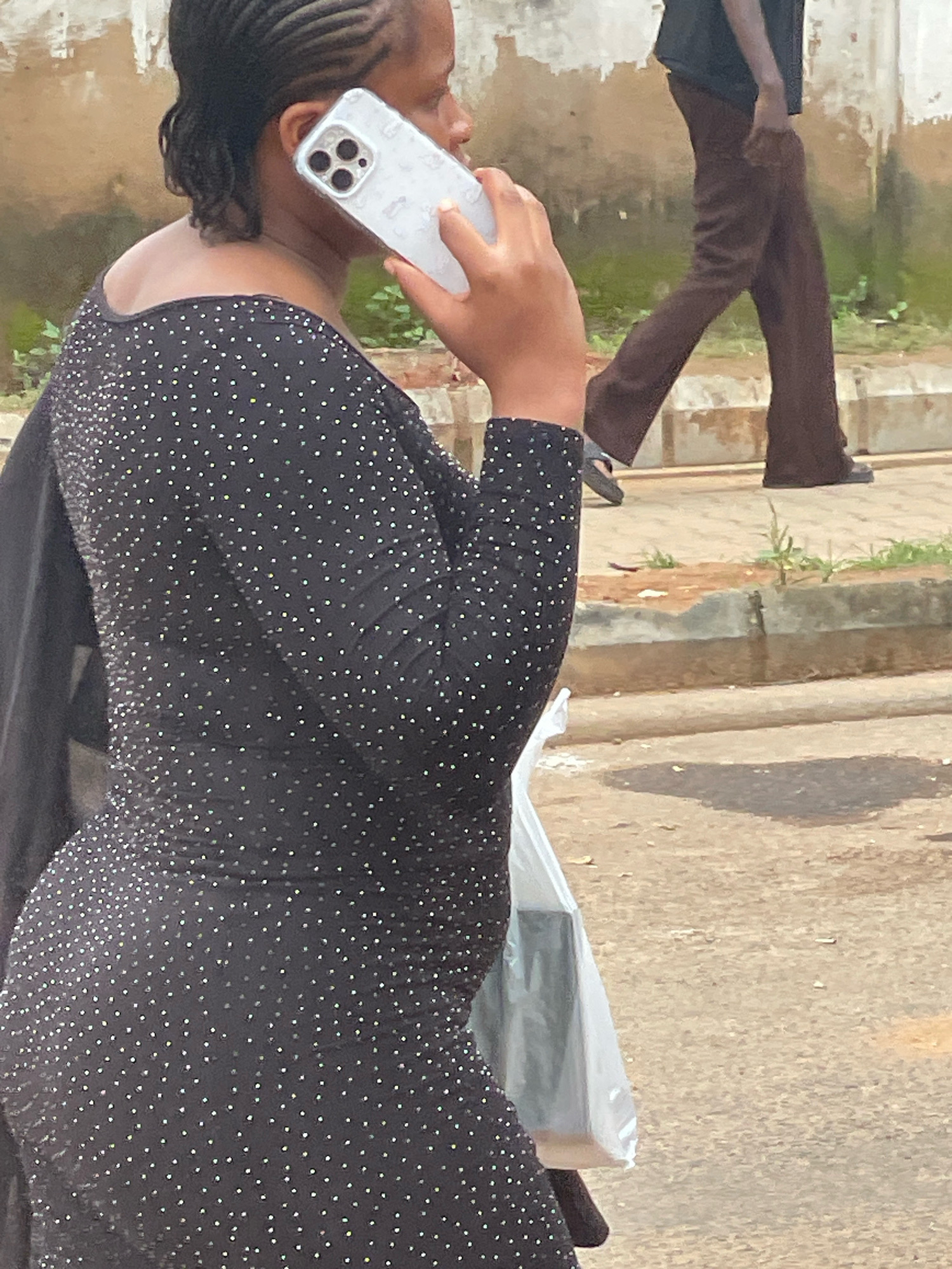 A woman with braided hair is on the phone, holding a white phone to her right ear. She is wearing a black dress adorned with numerous small, shiny studs. The woman is seen from the right side, from the shoulder down to her hip. Partially visible in the background, to the right of the woman, is a person wearing brown pants and a dark top, walking away from the camera. The scene appears to be outdoors, on a paved surface that could be a street or sidewalk, with some dirt and vegetation visible in the background. The environment suggests a casual urban setting in Abuja, Nigeria, possibly a public area. A white plastic bag is visible near the woman's hip.