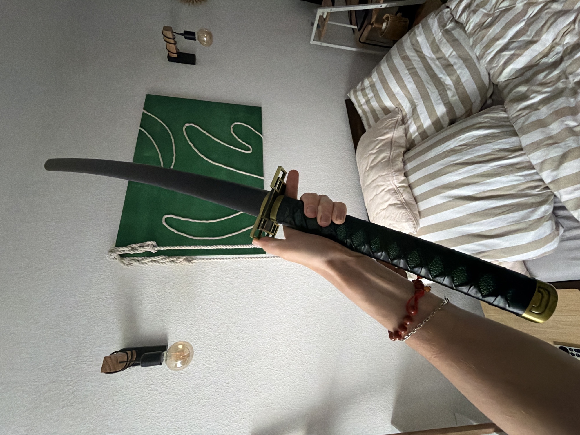 The image shows a person's arm holding a black sword with a green handle and a gold guard. The sword is positioned diagonally across the frame, with the tip pointing upwards and to the left. The background consists of a white textured wall with two wall-mounted lights and a green abstract painting. To the right of the painting, there is a shelving unit with some objects on it. Below the wall, a bed with striped bedding and pillows is visible. The overall scene appears to be an indoor setting, possibly a bedroom. There is no information in the image to suggest it is located in Herne, Germany.