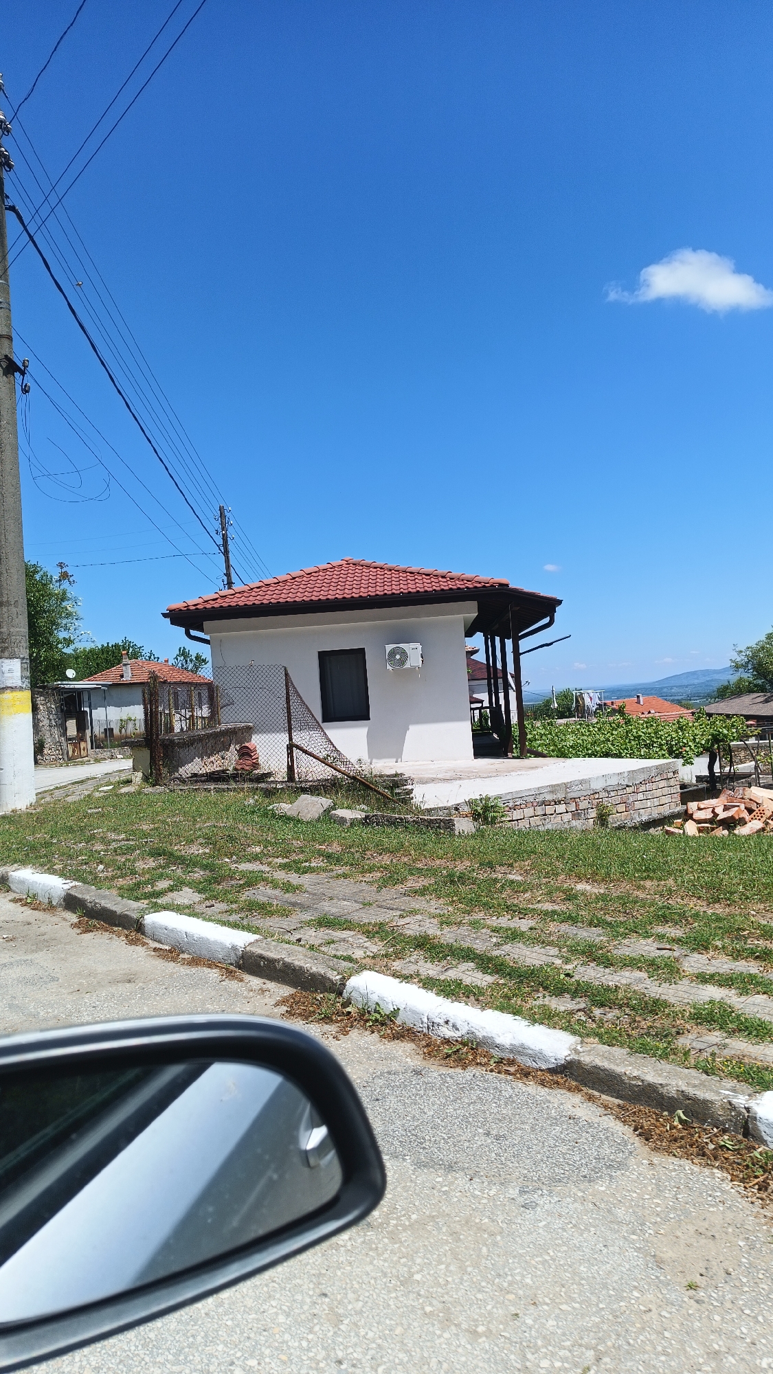 The scene is in Gornoslav, Bulgaria, showing a small, white building with a red tiled roof. The building features a window and an air conditioning unit mounted on its exterior wall. A partially collapsed chain-link fence borders the property. To the side, a bricked patio area is visible, along with some stacked bricks. The building is situated on a patch of grassy ground interspersed with paving stones. In the foreground, the side mirror of a car is visible, indicating the shot was taken from a vehicle. Overhead are power lines stretching across the sky, which is blue with a few scattered clouds. In the distance, rooftops of other buildings and some indistinct mountainous terrain can be seen.