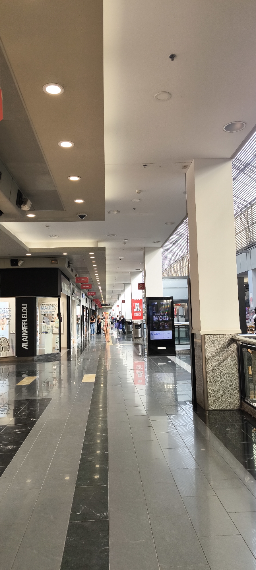 The image captures a well-lit, spacious shopping mall corridor, characterized by a clean, modern aesthetic. The scene is illuminated by numerous recessed circular lights in the ceiling, which vary between light beige and white panels. The floor is made of reflective tiles in alternating patterns of dark grey/black marble and lighter grey, creating a shiny surface that reflects the overhead lights and store displays.
Several people are visible in the distance, walking along the corridor, suggesting typical mall activity like shopping or commuting. One person in light-colored clothing is walking towards the camera in the mid-ground.
Notable elements include various shopfronts lining both sides of the hallway. On the left, a prominent black sign reads "ALAIN AFFLELOU," indicating an optician. Another store front has a sign that reads "MAGIC EXCLUSIVE" in white text on a black background, with "ENVIE DE STYLE!" and "VIVE LE STYLE!" (French for "Desire style!" and "Live the style!") visible on a poster. Further down the hall on the right, a red and white sign for "KFC" is clearly visible, with the slogan "PLANTANDO PODER" (Spanish for "Planting Power"). Beside the KFC sign is a tall, black digital advertising screen displaying text that includes "ÚNETE A WOB!" (Spanish for "Join WOB!").
The ceiling structure on the right side of the corridor features a slatted or louvred design, allowing some natural light to filter in, suggesting it is daytime. Security cameras are also visible on the ceiling. Some distant signs appear to have Arabic script alongside Latin characters, hinting at a multicultural environment or specific store offerings. The overall ambiance is bright, orderly, and functional.