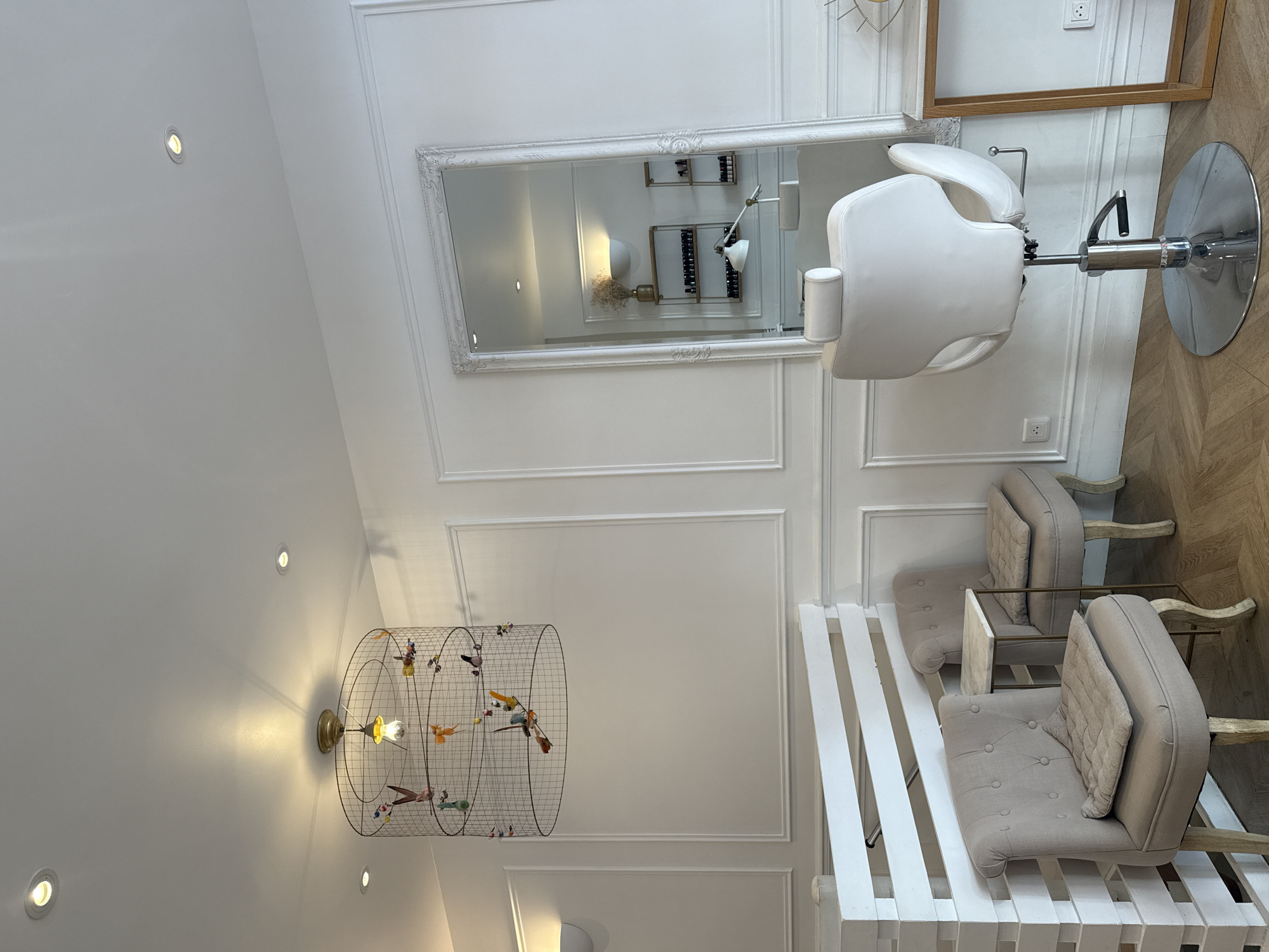 This is an interior photograph of a brightly lit salon, likely a nail salon or hair salon, in Paris, France. The walls are painted white and feature decorative molding. In the center of the room, a large ornate mirror is propped against the wall. To the left of the mirror, a white hydraulic styling chair with a chrome base sits on a wooden floor.
To the left of the styling chair, two upholstered chairs in a muted beige color are arranged near a white wooden partition. A small side table with a marble top is placed between them.
Hanging from the ceiling is a decorative light fixture that resembles a large birdcage, filled with colorful, decorative birds. The ceiling has recessed lighting.
Reflected in the mirror are shelves containing various bottles and a decorative vase with dried flowers, as well as a white desk lamp. The overall aesthetic is clean, modern, and elegant. There are no people visible in the image.