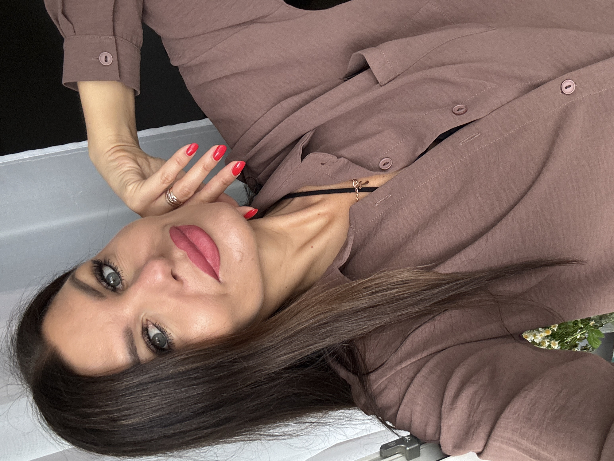 A medium shot, self-portrait photo taken from a low angle shows a woman with long dark hair, a light complexion, and blue eyes. She is wearing a mauve-colored collared shirt, unbuttoned at the top, revealing a black tank top underneath and a gold cross necklace. Her right hand is raised to her cheek, with bright red painted nails. Her lips are painted a matte pink. The background is out of focus, with a white curtain on the left and a dark wall on the right. There are no identifiable landmarks or objects in the image that would indicate a specific location within the city of Chaikovsky, Russia.