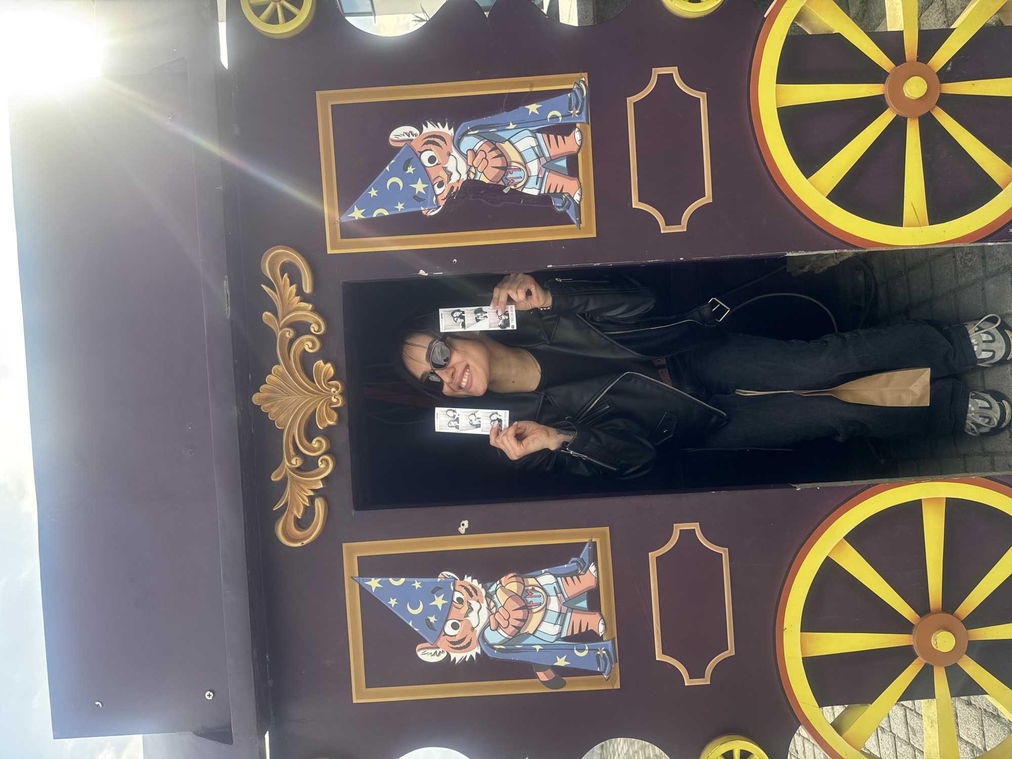A bright, daytime outdoor photo captures a person posing within a novelty carriage-like photo prop, likely in Tbilisi, Georgia. The main subject, a woman with dark hair and sunglasses, smiles broadly as she looks towards the camera. She is wearing a black leather jacket, dark jeans, and sneakers, and holds two photo booth strips in her hands.
The photo prop is a large, dark purple structure adorned with ornate gold scrollwork and features two prominent yellow wheels with red rims on either side. Incorporated into the prop are two decorative cutouts of a cartoon tiger character, each wearing a blue wizard hat and a blue robe with yellow stars and moons. The tiger at the top of the prop is depicted holding a staff, while the one at the bottom is shown from the waist up.
Strong sunlight causes a prominent lens flare in the upper left corner of the image, indicating a sunny day. The ground beneath the prop appears to be paved with light-colored stones. On one of the photo strips held by the woman, faint text reading "PHOTO" and a small logo are visible. The other strip clearly shows Korean characters, "인생네컷" (Insaengnecut), a popular Korean photo booth brand, suggesting the photo strips were recently taken from a photo booth. The overall scene depicts a cheerful moment of leisure and entertainment.