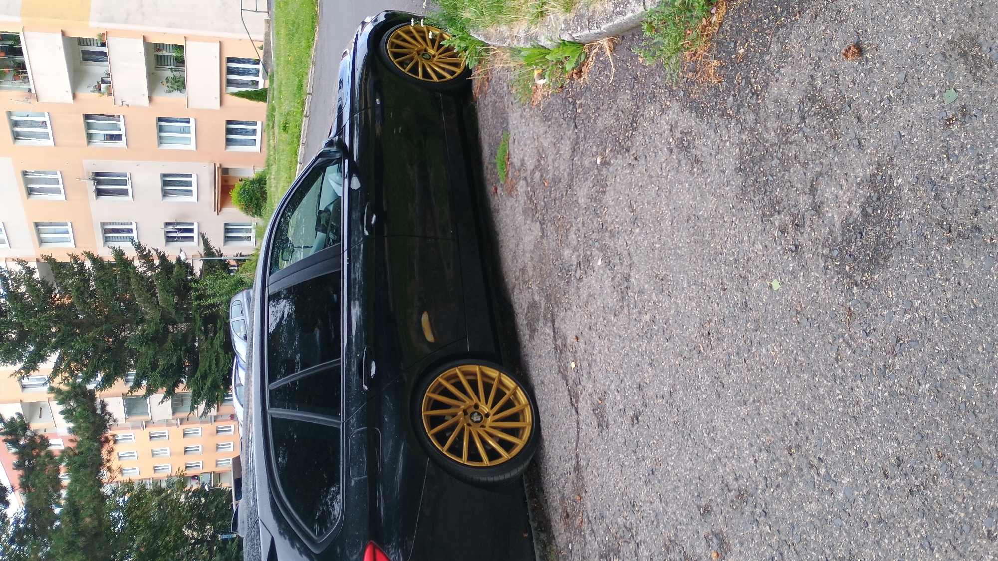 The image depicts the side view of a black station wagon parked on an asphalt surface. The car has distinctive gold-colored multi-spoke alloy wheels. In the background, a multi-story residential building with cream and peach-colored facades and numerous windows is visible, along with evergreen trees and some grass. The scene appears to be an urban residential area. The specific location is in the city of Sokolov, Czechia.