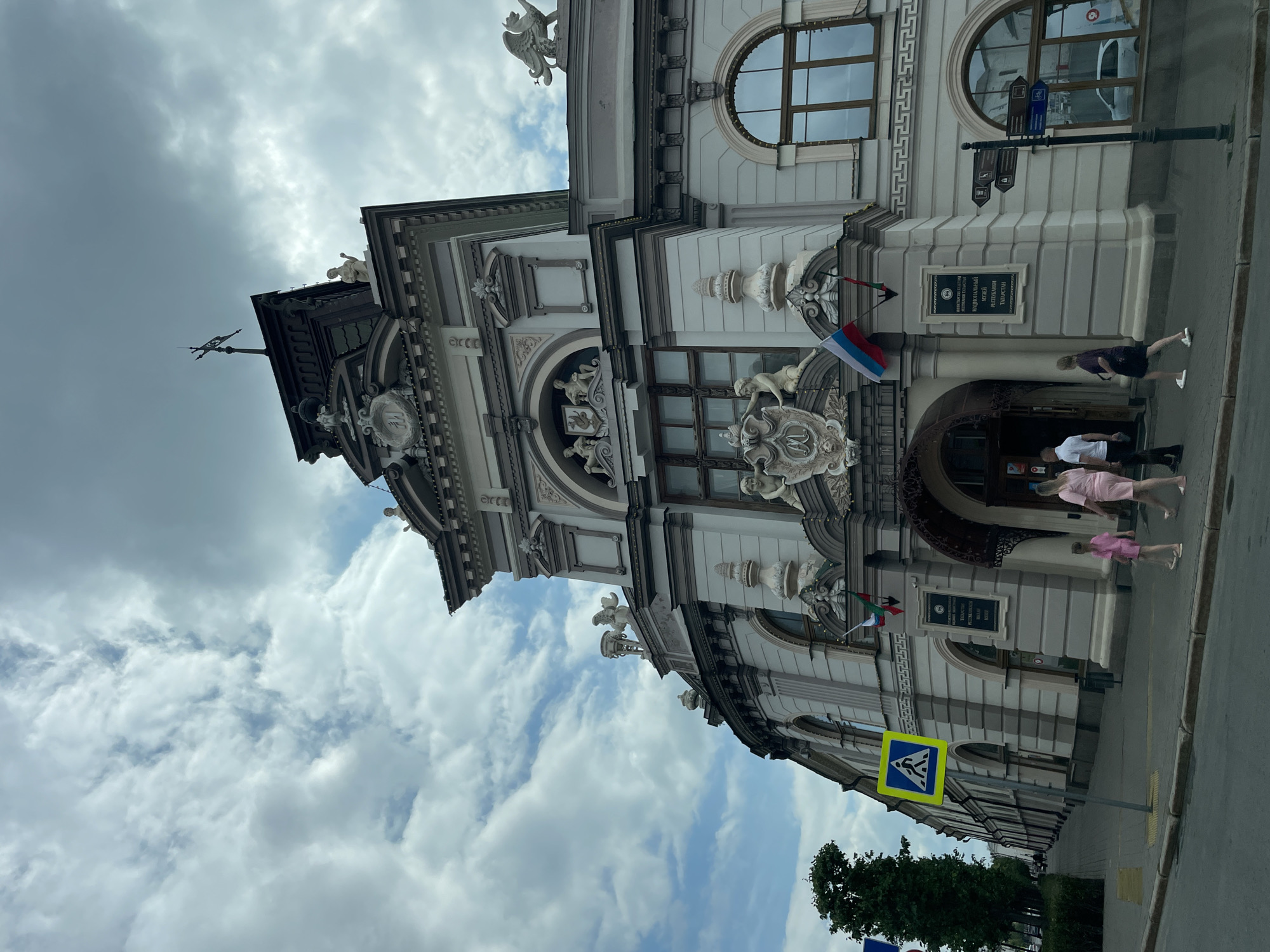The image shows the National Museum of the Republic of Tatarstan in Kazan, Russia. The museum is a grand, ornate building with neoclassical architecture, featuring columns, statues, and intricate carvings. The facade is light-colored with dark gray accents. In front of the museum, there are several people walking on the sidewalk. A pedestrian crossing sign is visible on the left side of the image. The sky is cloudy, with patches of blue peeking through. The overall impression is one of a historic and culturally significant landmark.
