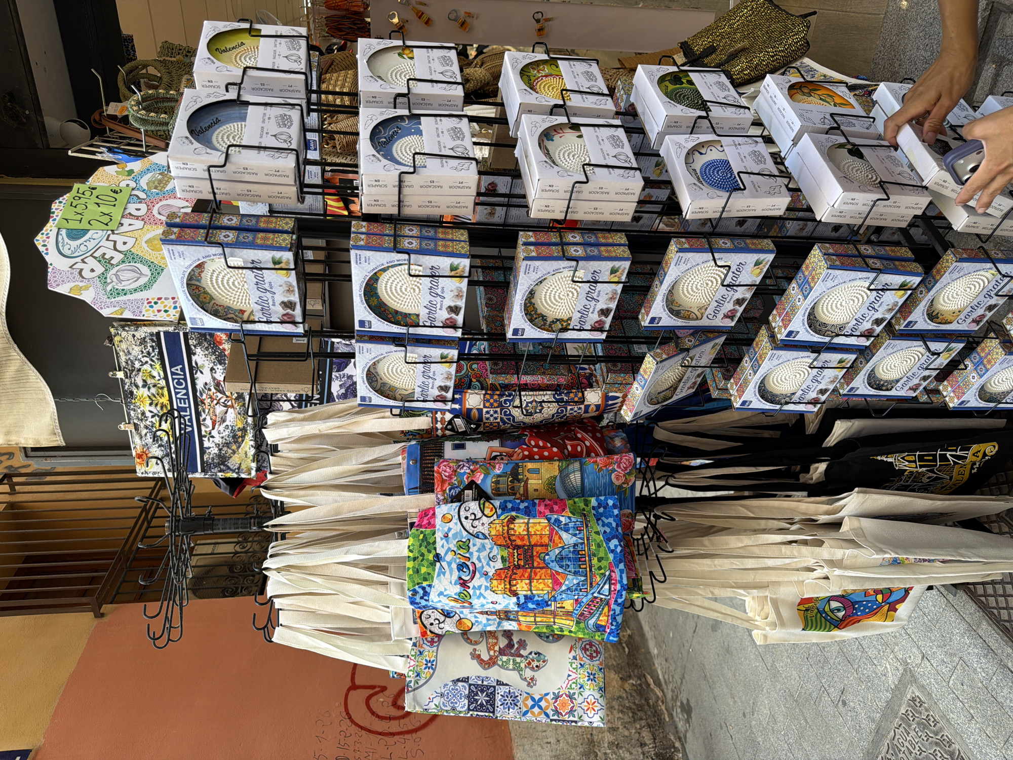 An outdoor market stall displays various merchandise for sale. In the foreground, a rack holds numerous beige canvas tote bags, some featuring colorful graphic prints of buildings, abstract art, and local landmarks, while others are plain. To the right of the tote bags, a black metal display rack is filled with numerous boxes of garlic graters, each box adorned with a picture of the grater and labeling in multiple languages. Some of these boxes are stacked three high, while others are displayed individually. In the background, more items are visible, including patterned fabric and a display of woven baskets. A brick wall painted orange and a metal grate are also visible. A hand is reaching for a garlic grater box.