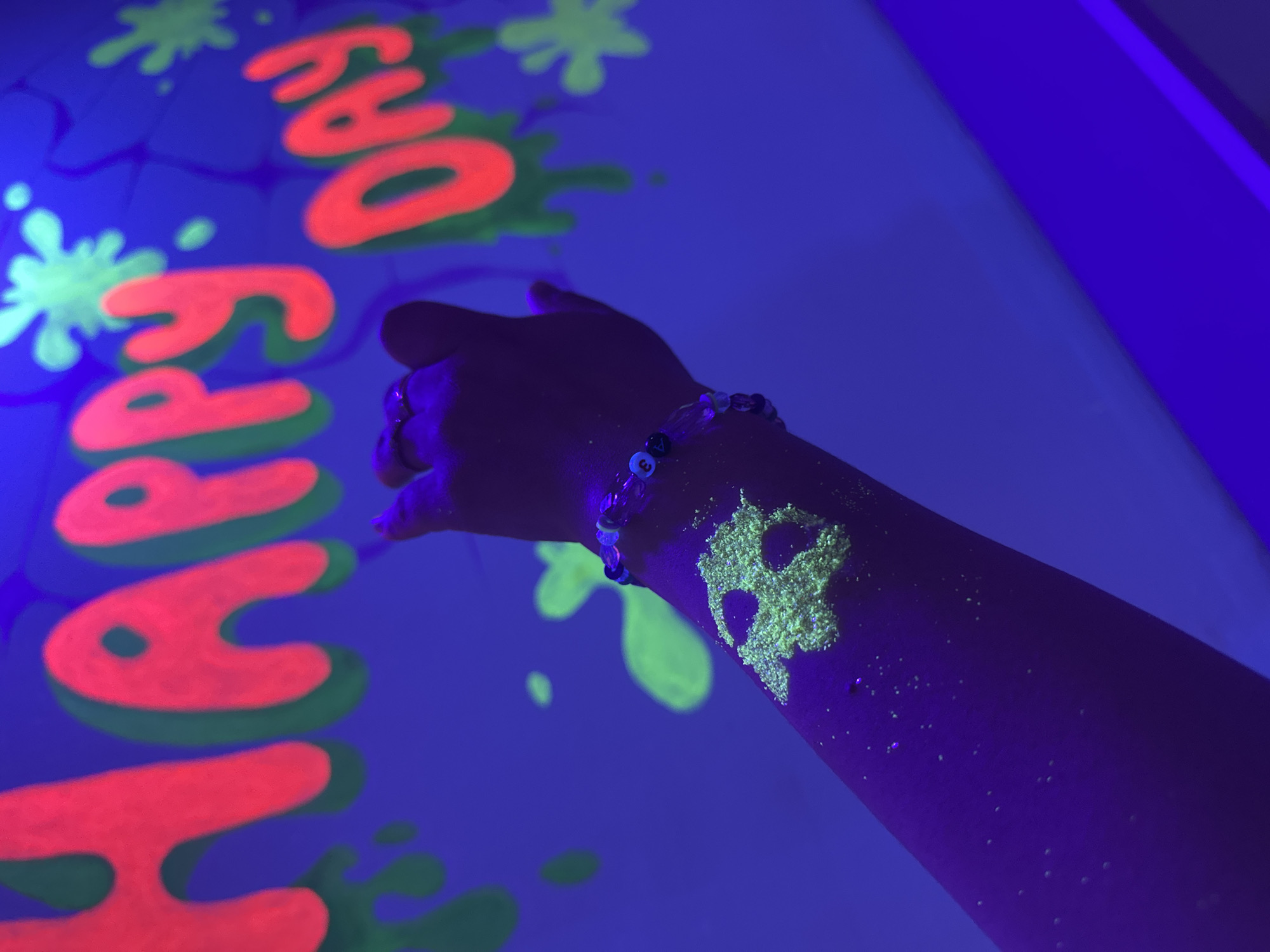 A photograph taken under ultraviolet light shows a person's right arm and hand extended towards a blue surface. The surface displays the text "Happy Day" in red with green outlines, along with green splatter-like shapes. The person wears a beaded bracelet. There is a lime green design, possibly a skull, on the person's wrist. The rest of the arm is illuminated with what appears to be small particles, perhaps glitter. The location of the city is Unknown, Unknown.