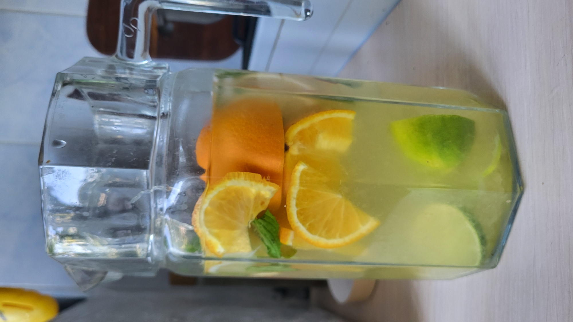 This media file captures a close-up, top-down view of a glass beverage dispenser filled with a refreshing drink. The dispenser contains slices of oranges, limes, and a few sprigs of mint, infusing the yellow liquid with vibrant colors and likely a citrusy flavor. The dispenser itself is made of clear glass and features a silver spigot for serving. The lighting suggests it's daytime, possibly indoors given the clean, light-colored surface the dispenser rests on, which appears to be a countertop or table. In the background, a hint of a tiled wall and a wooden surface can be seen, suggesting a kitchen or dining area. The overall impression is one of a cool, inviting beverage prepared for enjoyment. There is no visible text in the image.