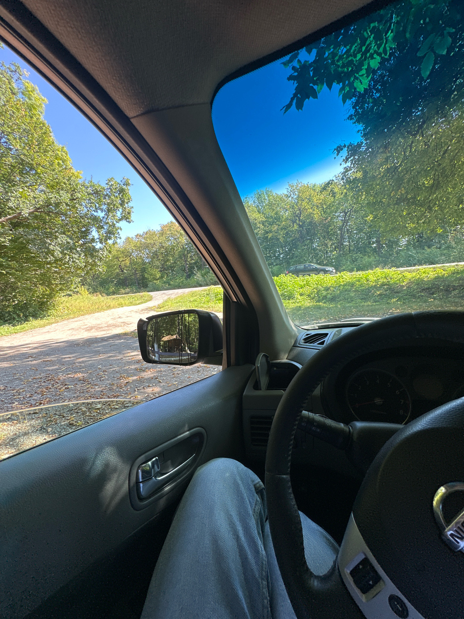 The image depicts a point-of-view shot from inside a Nissan vehicle, looking out towards a rural road. The perspective is from the driver's seat, with the steering wheel and part of the dashboard visible in the foreground. The driver's legs, clad in light blue jeans, are also visible.
The exterior environment is characterized by a dirt road winding through a wooded area with lush green trees under a clear blue sky. Sunlight filters through the leaves, casting dappled shadows. In the distance, another vehicle, a dark SUV, is visible on a paved road to the right. A side mirror on the car reflects a small wooden cabin or structure amidst the trees. The scene suggests a drive through a natural, possibly remote, landscape. There is no information in the image to suggest the location is the city of Shatava, Ukraine.