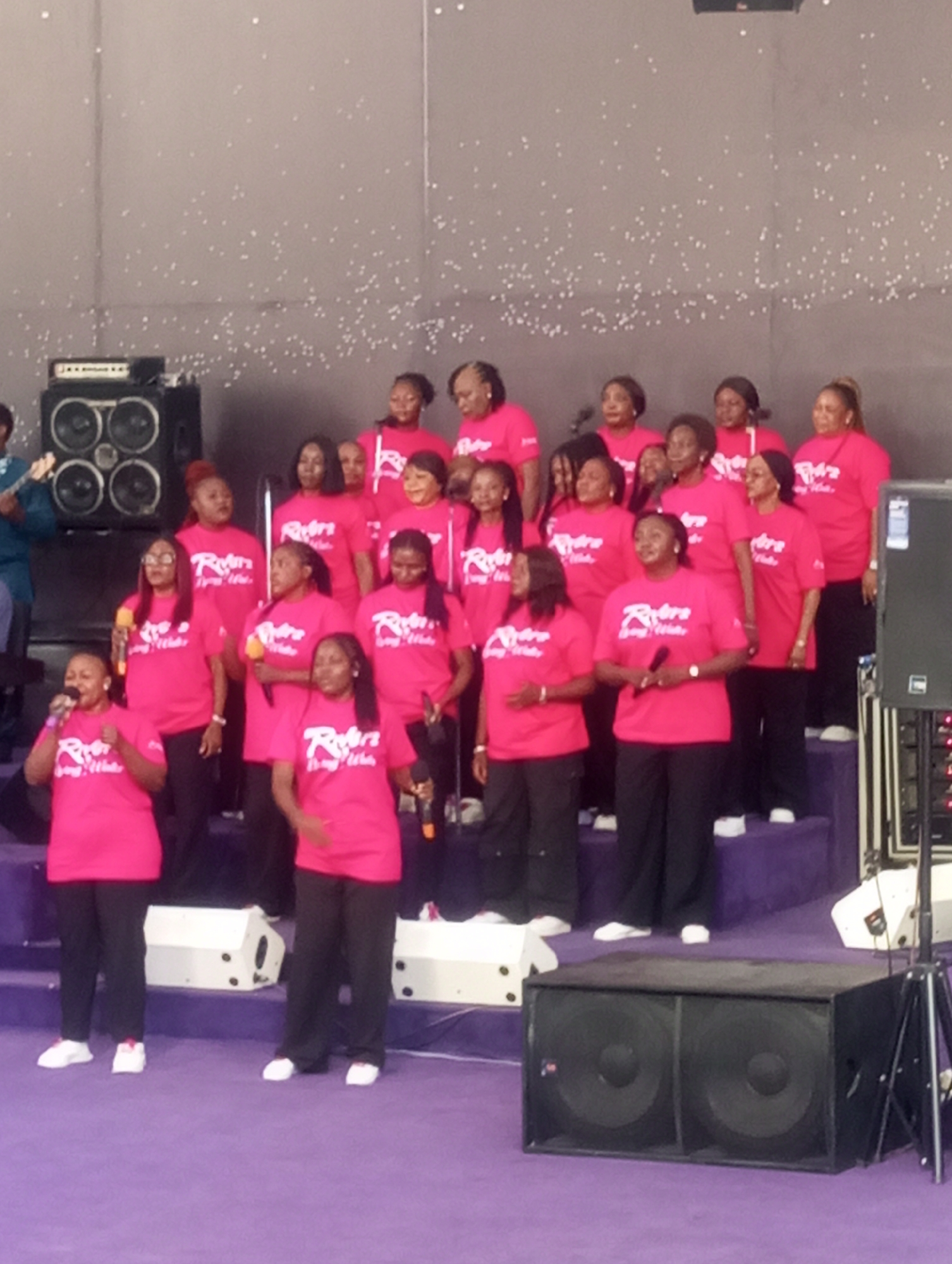 On a stage covered in purple carpet in Gura Top, Nigeria, a large choir of African women wearing matching bright pink T-shirts with a logo and black pants sing into microphones. A man plays guitar to their left. Speakers and musical equipment are visible on the stage. A textured gray wall forms the backdrop.