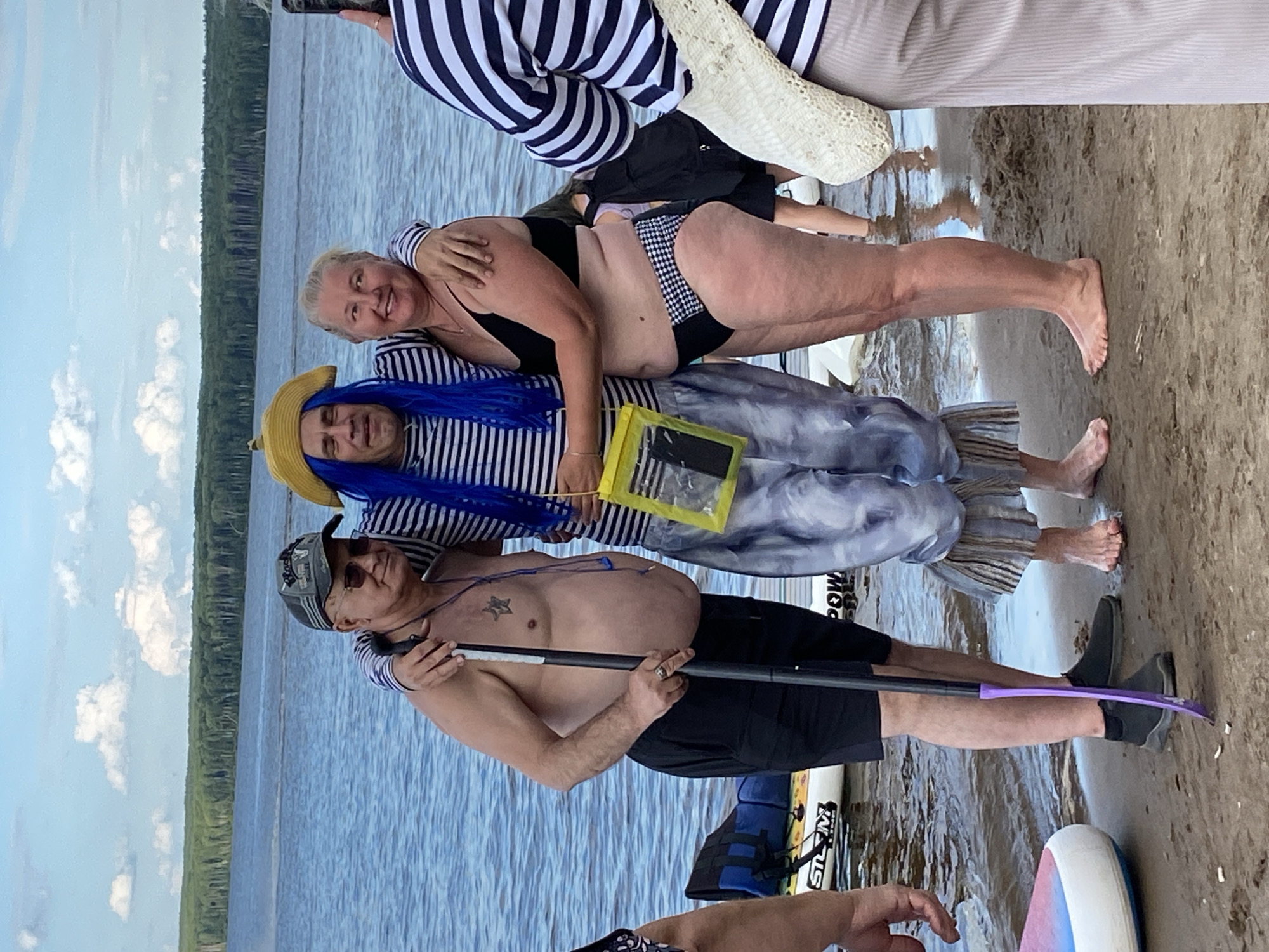 This media file depicts a group of people on a sandy beach with a body of water in the background. The scene is oriented vertically, as if the camera was rotated.
The primary subjects are three people. The person in the front is a man with a bare chest, wearing black shorts and a baseball cap. He is holding a paddle. Behind him and to his left, a woman with blonde hair is smiling and wearing a black swimsuit. Next to her and further back, a person in a blue wig and a striped shirt is also visible. Several other individuals are partially visible in the background, some wearing striped clothing.
Objects present include the paddle held by the man, a yellow waterproof pouch, and what appears to be a paddleboard or inflatable boat near the man's feet. In the background, a dense line of trees borders the water, suggesting a natural setting.
The time of day appears to be daytime, given the lighting and the presence of clouds in the sky. The weather seems pleasant, with a clear sky and calm water. The presence of people in swimwear and near water suggests a recreational activity, possibly paddleboarding or enjoying a day at the beach.
No text is clearly legible in the image. The overall mood appears relaxed and cheerful, with smiles visible on some of the subjects' faces. The location context provided is Chaikovsky, Russia.