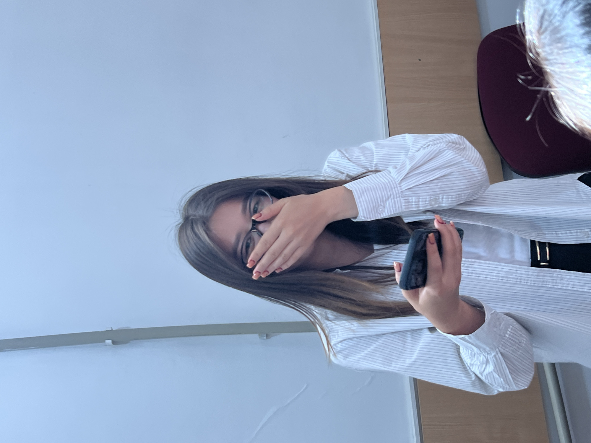 A young woman with long brown hair and glasses is seated indoors, holding a black smartphone in her right hand. She is covering her mouth with her left hand, suggesting surprise or amusement. She is wearing a white striped button-down shirt over a white tank top and a black belt. The background appears to be a classroom or meeting room, with a plain white wall and a wooden accent wall. The location is identified as Kadamjay, Kyrgyzstan.