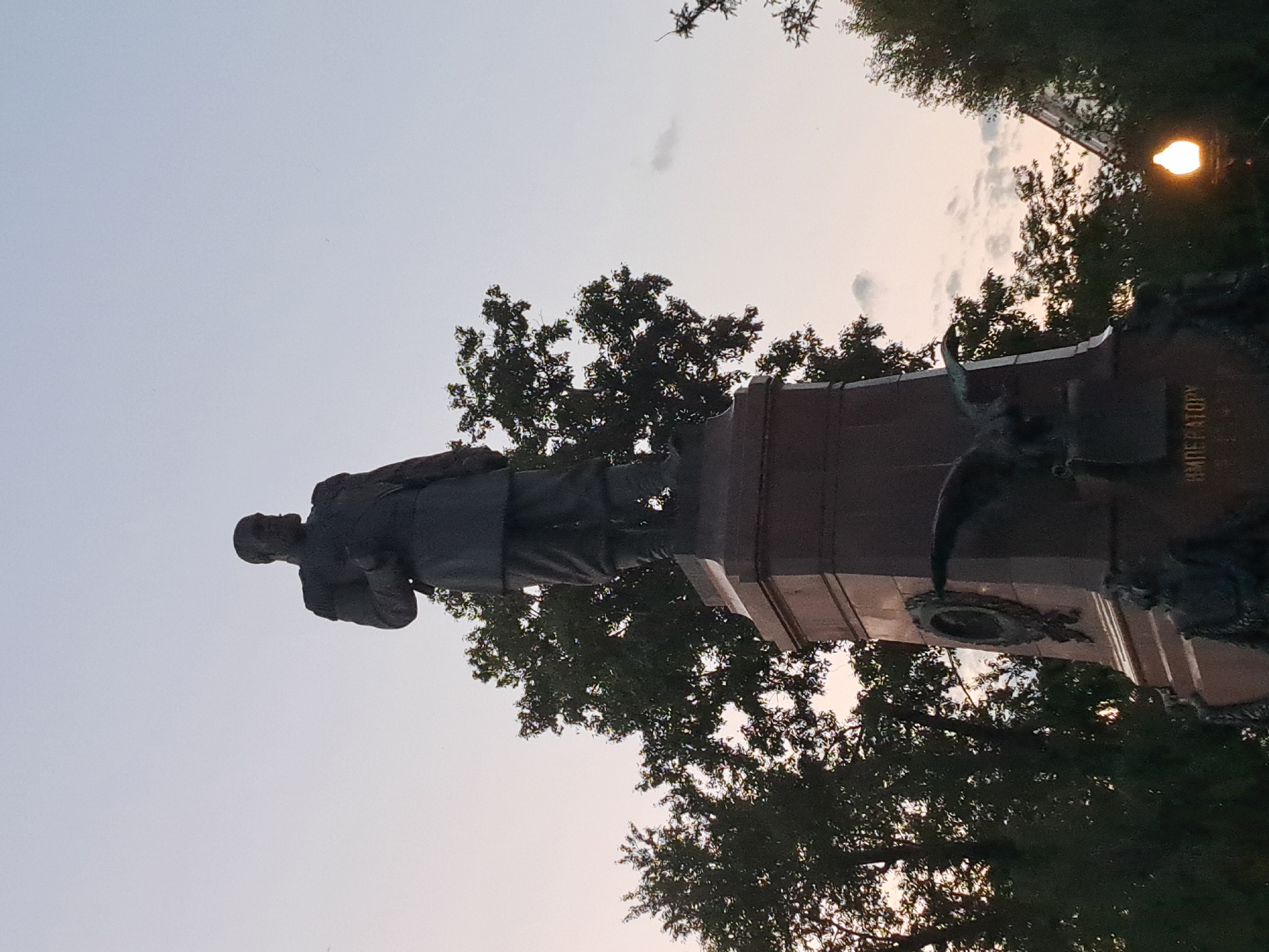 The image shows a statue of a man in uniform standing on a large pedestal. The statue is dark in color, and the pedestal is made of stone. There are trees around the statue, and the sky is visible in the background. A street lamp is also visible in the lower right corner of the image. The inscription on the pedestal reads "ИМПЕРАТОРУ," which translates to "To the Emperor." This is a statue of Emperor Alexander III, located in Irkutsk, Russia. The statue was erected in 1909.