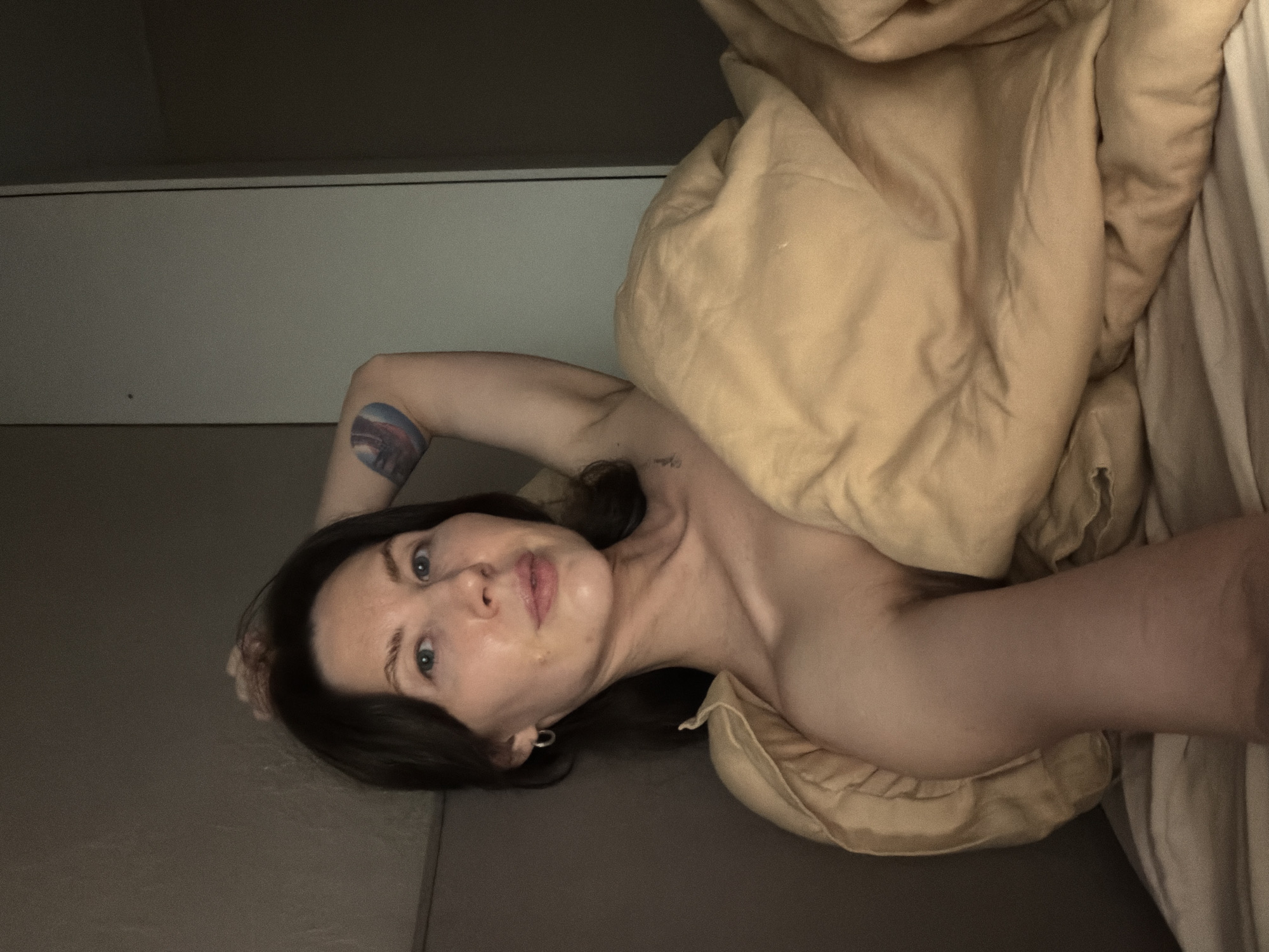 A woman is lying in a bed covered in a tan duvet and white and gray sheets. She has dark hair, fair skin, and blue eyes. A tattoo is visible on her left arm. The environment appears to be a bedroom with white walls. There is no evidence to indicate this image was taken in Perm, Russia.