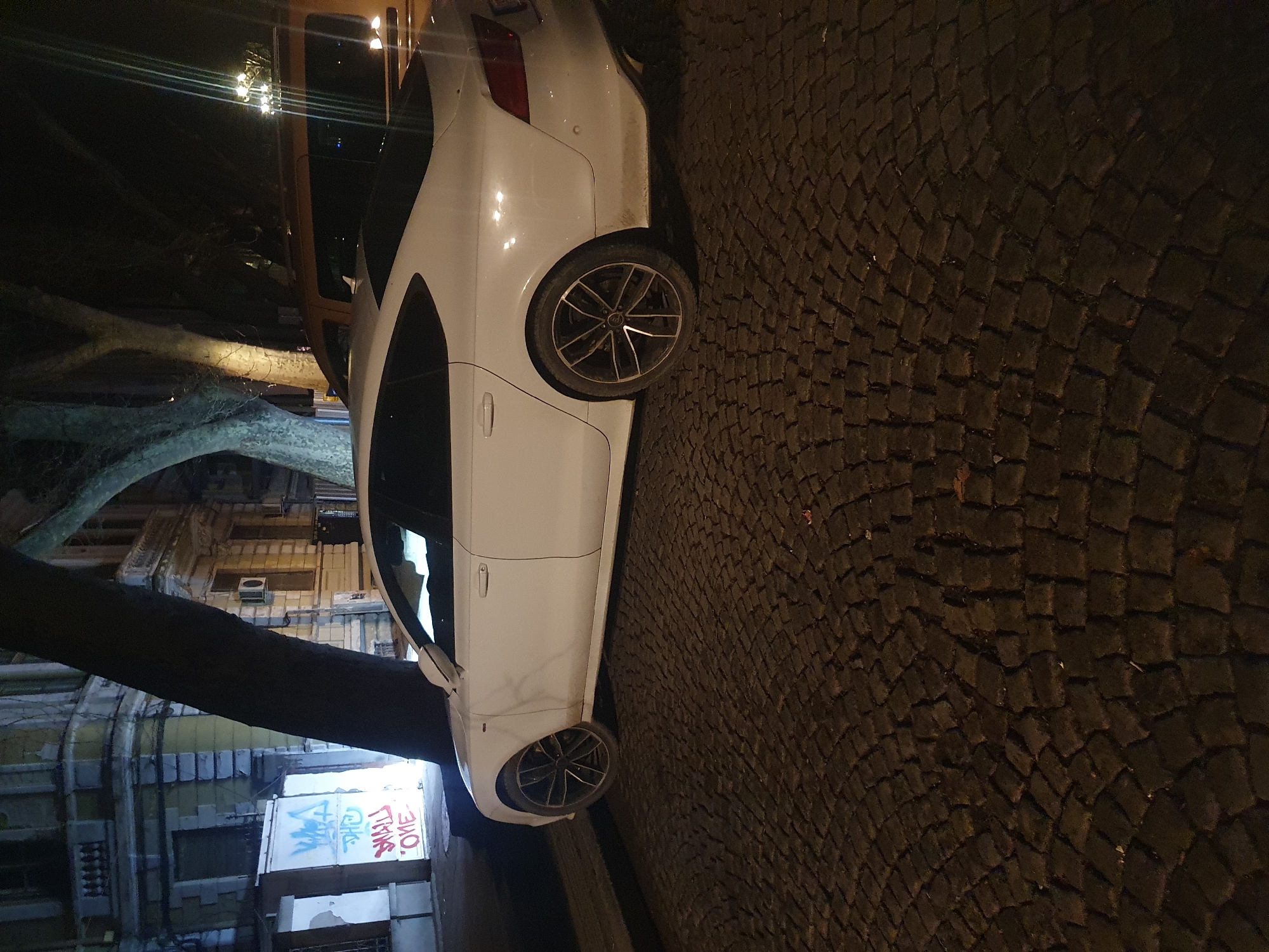 The image shows a white car parked on a cobblestone street. Above the car, a tree branch extends across the frame, and a building facade with graffiti is visible in the background. The scene appears to be illuminated by street lighting. Based on the building style and the cobblestone street, the location is likely in an older part of a European city. It is not possible to confirm the location is Plovdiv, Bulgaria without further information.