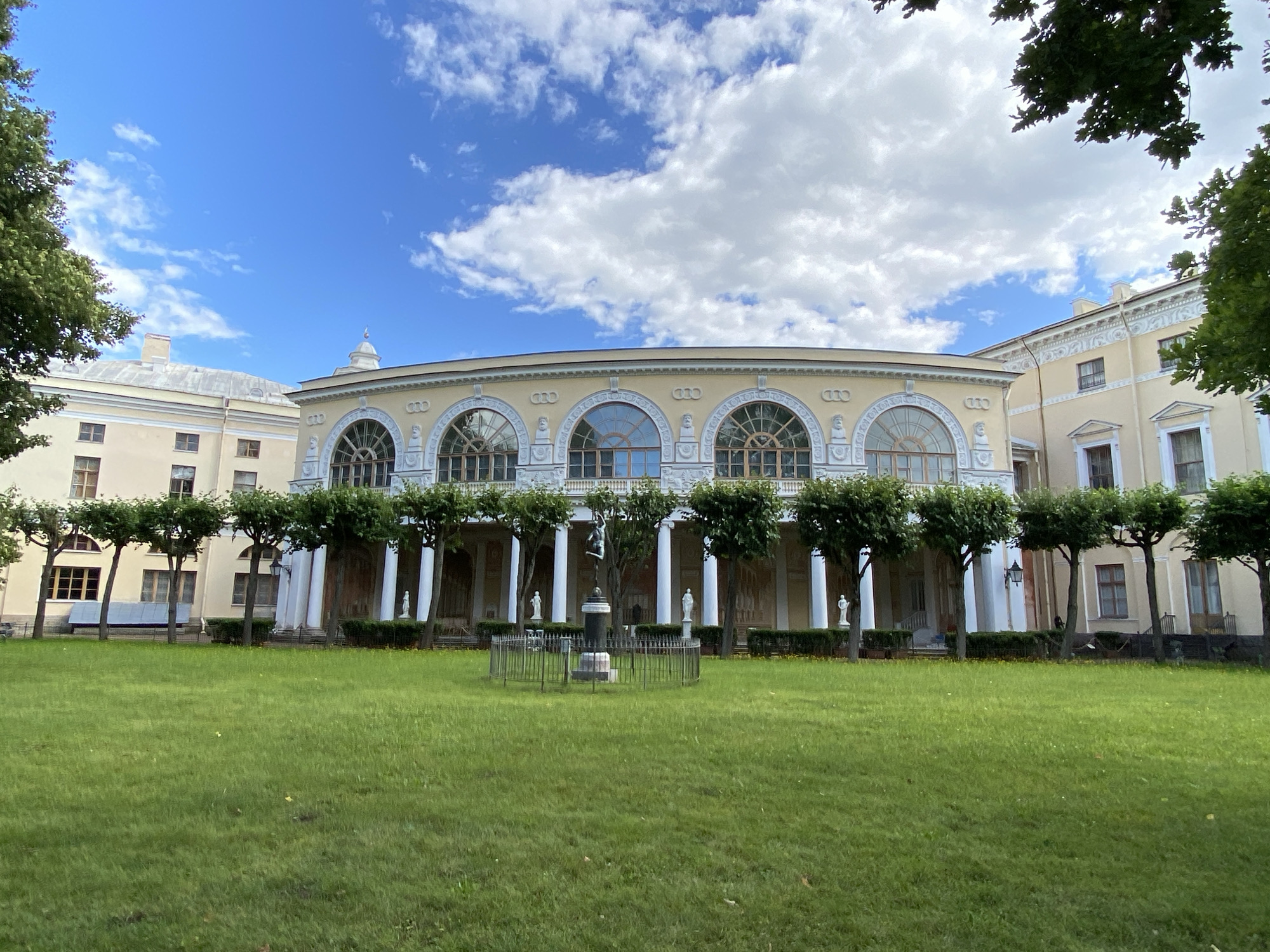 The image captures a frontal view of the Pavlovsk Palace ensemble, specifically the colonnade of the Rossi Pavilion, on a bright, partly cloudy day. The palace, painted in a pale yellow, features a grand, curved facade adorned with a series of large, arched windows framed by elaborate stucco work. A prominent colonnade with tall, white columns supports the upper level of the building. In front of the palace, a well-maintained lawn stretches out, leading to a central ornamental fountain surrounded by a small, black metal fence. Several manicured trees, pruned into rounded shapes, are planted in a row along the edge of the lawn, separating it from the palace. Statues are visible on pedestals behind the trees and near the palace entrance. The sky above is a vivid blue, with scattered white clouds. Lush green trees frame the upper right corner of the image, suggesting a park-like setting.