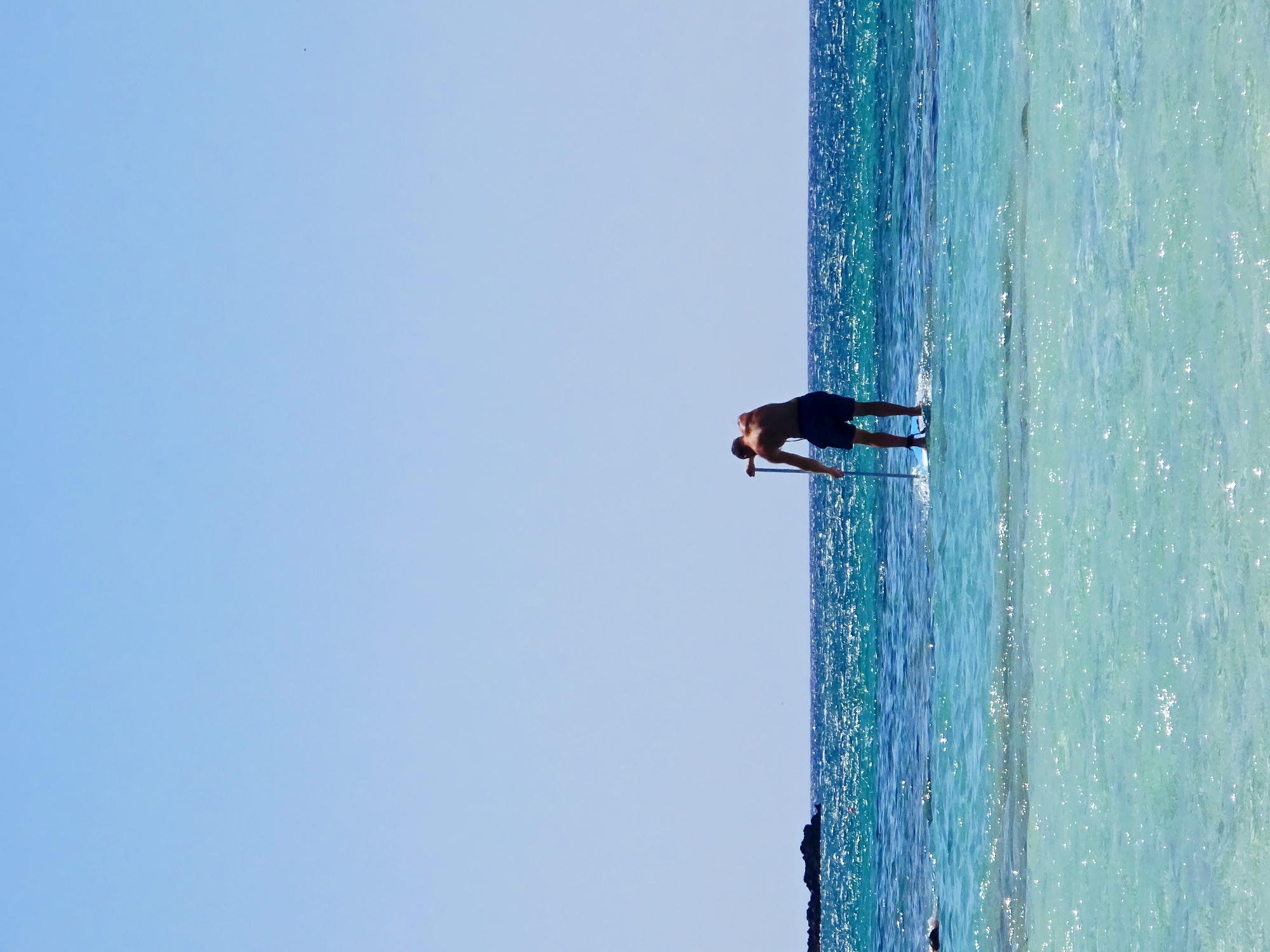 A person is stand-up paddleboarding in the clear turquoise waters of Greece. The individual, seen from behind, is wearing dark swim trunks and is holding a paddle. The water is calm with small waves and sunlight glinting off the surface. In the distance, a rocky island is visible. The sky is a clear, bright blue.