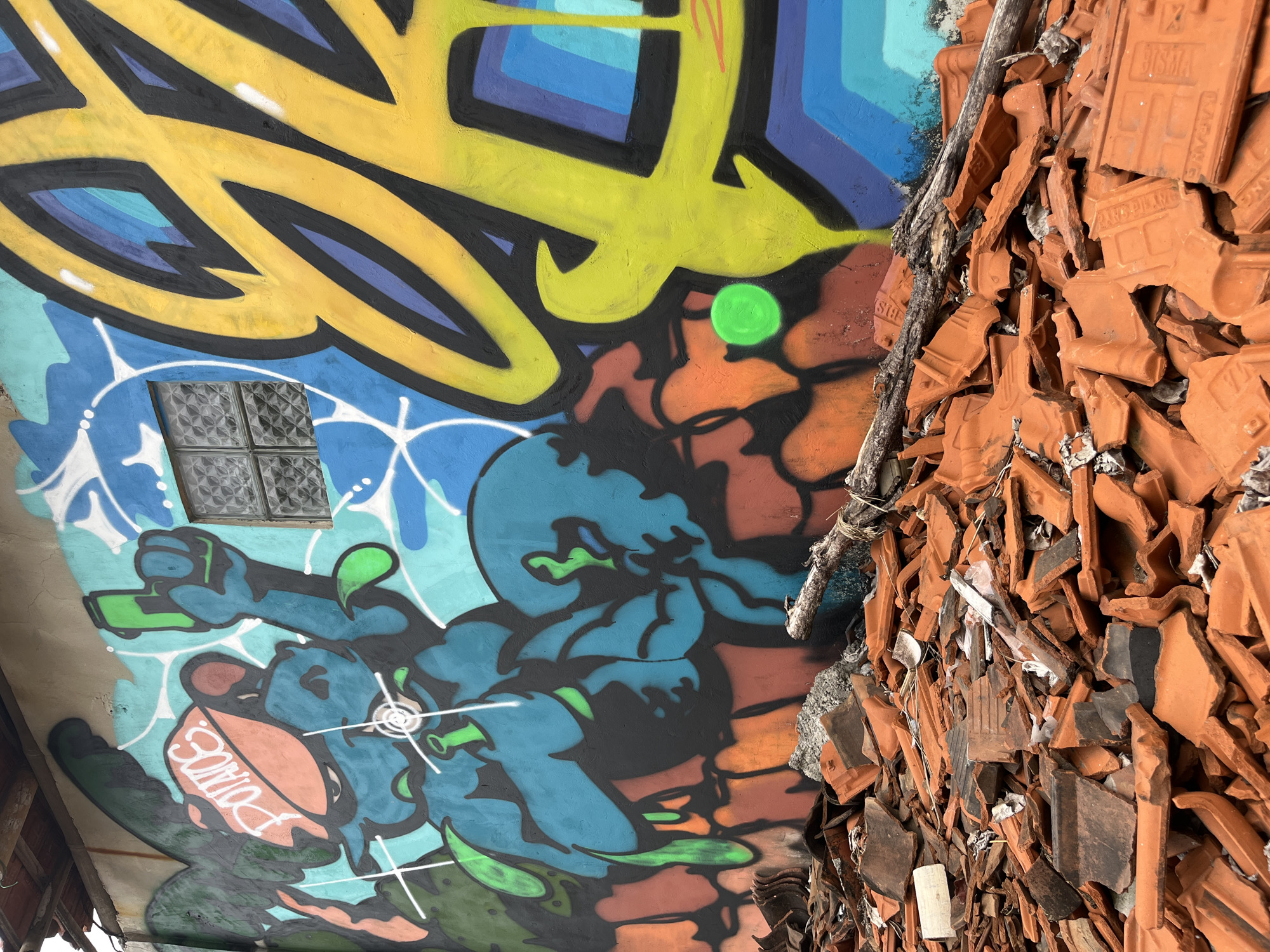 In Kuta Selatan, Indonesia, a close-up, low-angle shot captures a vibrant graffiti-adorned wall juxtaposed with a pile of broken terracotta roof tiles in the foreground. The graffiti features a cartoonish blue character wearing an orange cap with the word "POTATOS" in white. This character holds a green gun-like object, and crosshairs are visible on its chest. To the right, large yellow graffiti lettering dominates, with black outlines and purple and blue accents. A small, frosted glass window is set into the wall between the blue character and the yellow lettering. A green circle is painted on the wall to the right of the blue character. The broken roof tiles, in various shades of orange and brown, are scattered across the ground, some appearing partially covered by debris. A weathered wooden branch lies across the pile of tiles. The overall scene suggests a location undergoing demolition or renovation, with the colorful street art a stark contrast to the surrounding disarray.