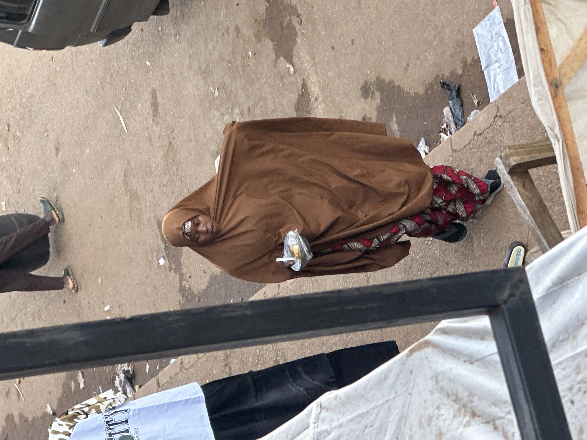 In Bukuru, Nigeria, a woman wearing a brown hijab and a red and black patterned skirt stands on a dirt path. She holds a small white bowl containing food in her right hand and a spoon in her left. The ground is littered with debris, including plastic bags and wrappers. A dark-colored van is parked to the right of the frame. In the background, another person, dressed in a light-colored shirt with a camouflage pattern and black pants, stands on the left side of the image. The scene appears to be an outdoor market or street.