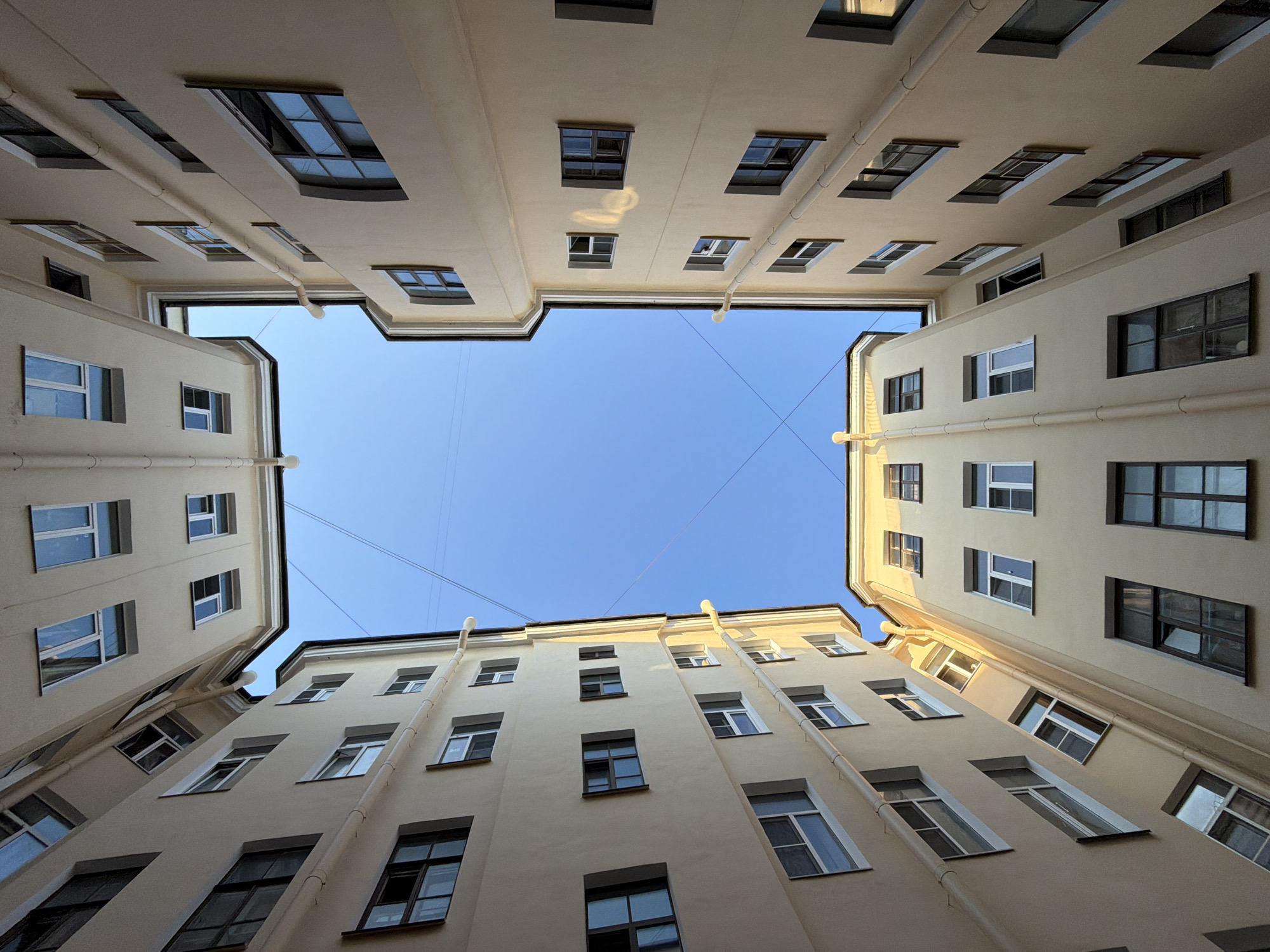 This is a low-angle, upward-looking photograph of a courtyard in Saint Petersburg, Russia, with buildings on all four sides. The buildings are tall, multi-story structures with beige facades and numerous windows. Some windows are open, while others are closed. There are downspouts running vertically along the exterior of the buildings. The sky above is a clear, bright blue, with a few thin wires crisscrossing it. The perspective of the photo creates a feeling of looking up into a narrow canyon of architecture. The scene is devoid of people, vehicles, or any other obvious signs of life, suggesting a quiet moment or an empty public space. The architecture appears to be of a late 19th or early 20th-century style, common in St. Petersburg. The lighting suggests it is daytime, possibly late afternoon due to the angle of the sunlight highlighting some of the building surfaces.