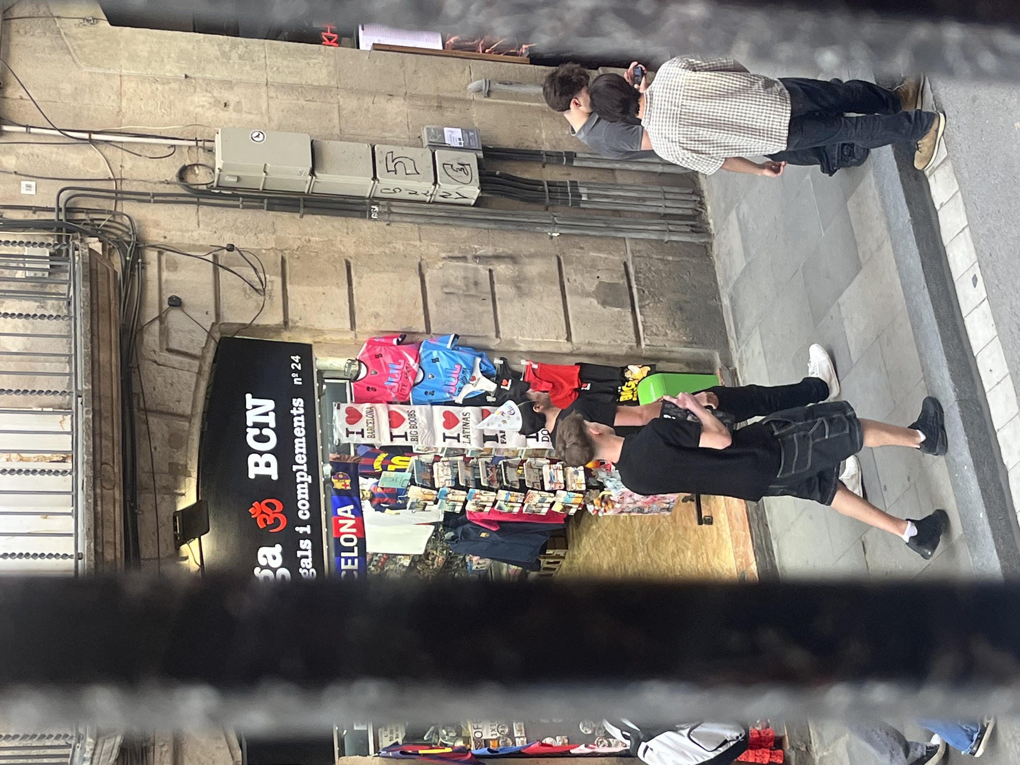 This image captures a street scene in Barcelona, Spain, featuring a souvenir shop and several pedestrians. The photo is rotated 90 degrees clockwise from its original orientation.
The setting appears to be a narrow pedestrian street or alleyway, characterized by old stone or concrete walls. Numerous electrical conduits and grey utility boxes are visible on the wall, indicating urban infrastructure. The ground is paved with light grey tiles.
On the left side, the entrance to a shop is prominently displayed, filled with merchandise. A black sign above the entrance reads "Sa ॐ BCN" (likely "Casa BCN", with an Om symbol) and "regals i complements nº 24", signifying a shop for gifts and accessories at address number 24. The shop's display racks are laden with various t-shirts and souvenirs. Visible t-shirt slogans include "I ❤️ BARCELONA", "I ❤️ BIG BOOBS", and "I ❤️ LATINAS". A blue banner also shows part of the word "CELONA".
Four individuals are present. Two are seen on the right, standing near the wall; one person, wearing a light plaid shirt over a dark t-shirt and dark pants, is looking at a mobile phone. Another person is partially obscured next to them. On the left, near the shop entrance, two more individuals are visible. One wears a black t-shirt and dark shorts, appearing to be observing the shop's display. The other is mostly out of frame.
The scene depicts a typical daytime urban environment, with people casually browsing or passing through a commercial area. The text on the merchandise and shop sign directly indicates the location as Barcelona.