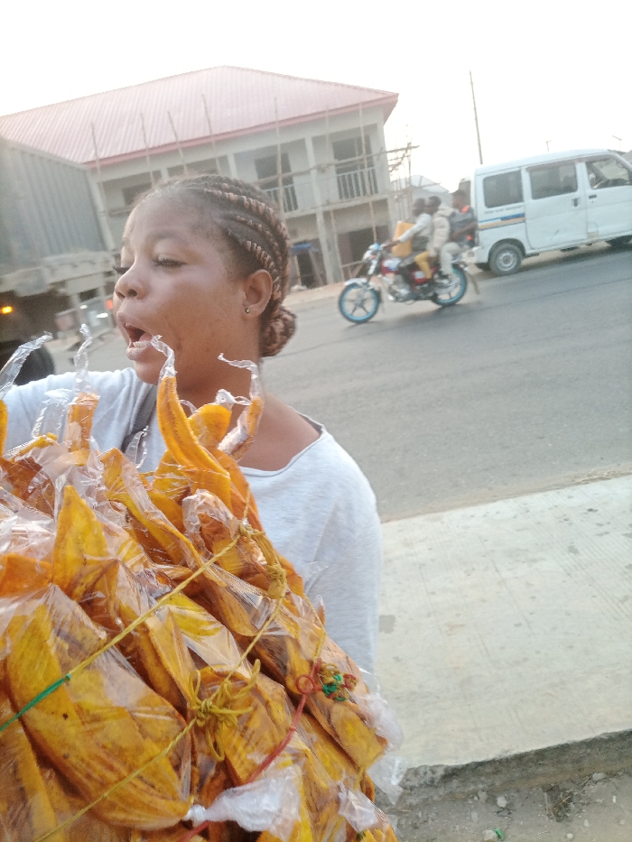 The image captures a street scene in Obehie, Nigeria. In the foreground, a young adult female with braided hair, wearing a light grey shirt, is positioned with her mouth wide open, possibly mid-yawn or speaking. She carries a substantial bundle of individually packaged, elongated yellowish-orange items, likely fried snacks, encased in clear plastic bags and secured with multicolored strings (yellow, red, green). Behind her, a paved road is visible. On the road, a white panel van is parked or moving slowly on the right, and a silver motorcycle with blue wheel accents carries two male occupants, one of whom has a yellow package on his back, in motion to the left of the van. A large, dark truck is partially visible on the far left. In the background, a two-story building under construction, featuring a red corrugated metal roof and exposed concrete walls with wooden scaffolding, stands beneath a bright sky. A concrete sidewalk or curb occupies the immediate foreground.