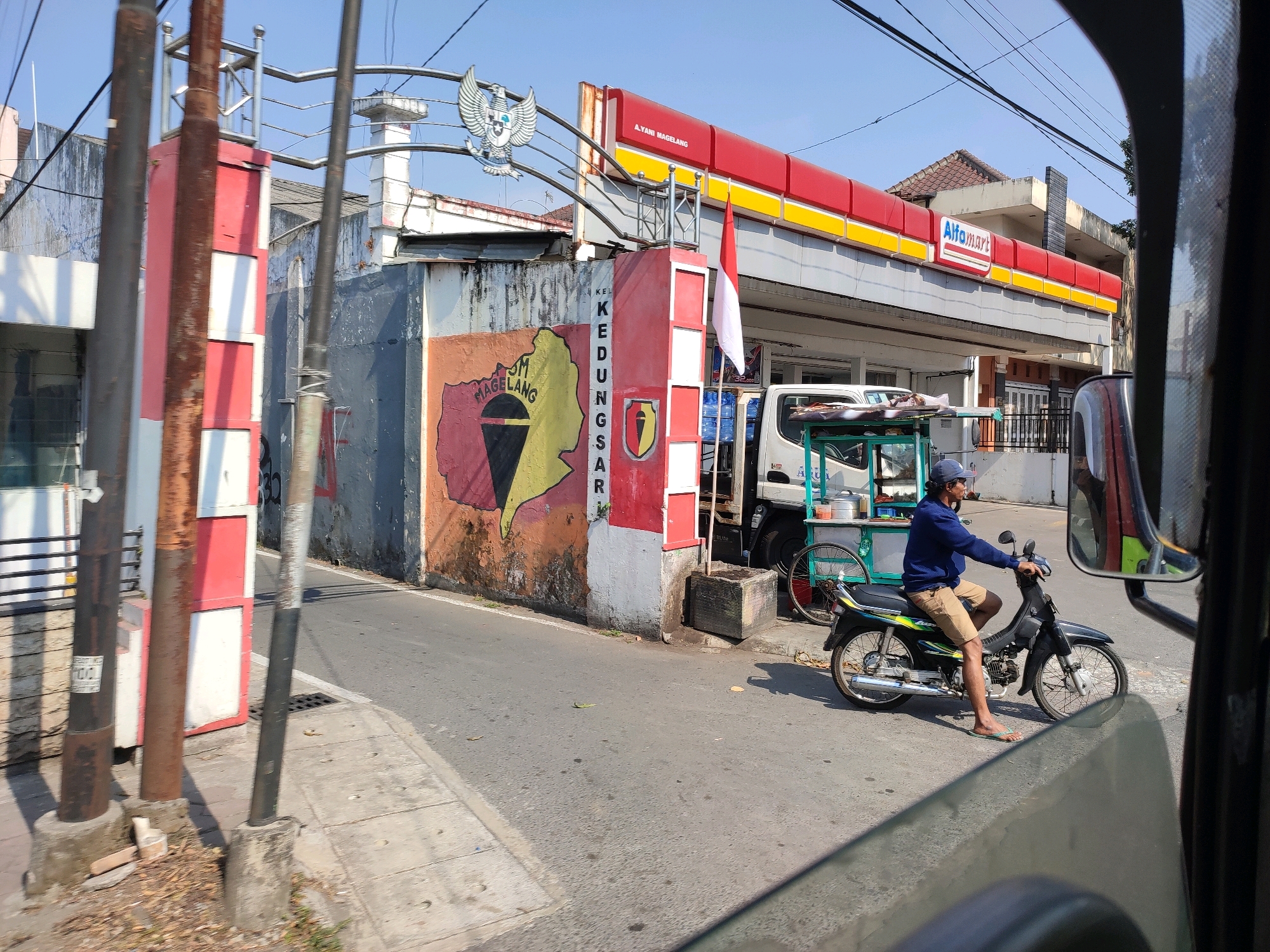 The image depicts a street scene in Magelang, Indonesia, likely near a main thoroughfare. In the foreground, a motorcycle rider, wearing a blue long-sleeved shirt, tan shorts, and a dark blue cap, is stopped on a paved road. The motorcycle is black with green and yellow accents and is facing left. To the left of the rider, a white truck with a green cart attached is parked.
Behind the motorcycle, a building with a red and yellow facade, bearing the "Alfamart" logo, is visible. The building appears to be a convenience store. To the left of the Alfamart, a large, weathered concrete wall is adorned with a colorful mural, depicting a map-like design with red, yellow, and black sections. A prominent red and white Indonesian flag is attached to a pole on the wall. Above the wall, a metal structure with an eagle emblem is present.
The environment is urban, with a clear blue sky overhead. Power lines are strung across the sky. The street is asphalt, with a concrete sidewalk on the left side. The overall impression is a typical street scene in an Indonesian city on a bright, sunny day.
