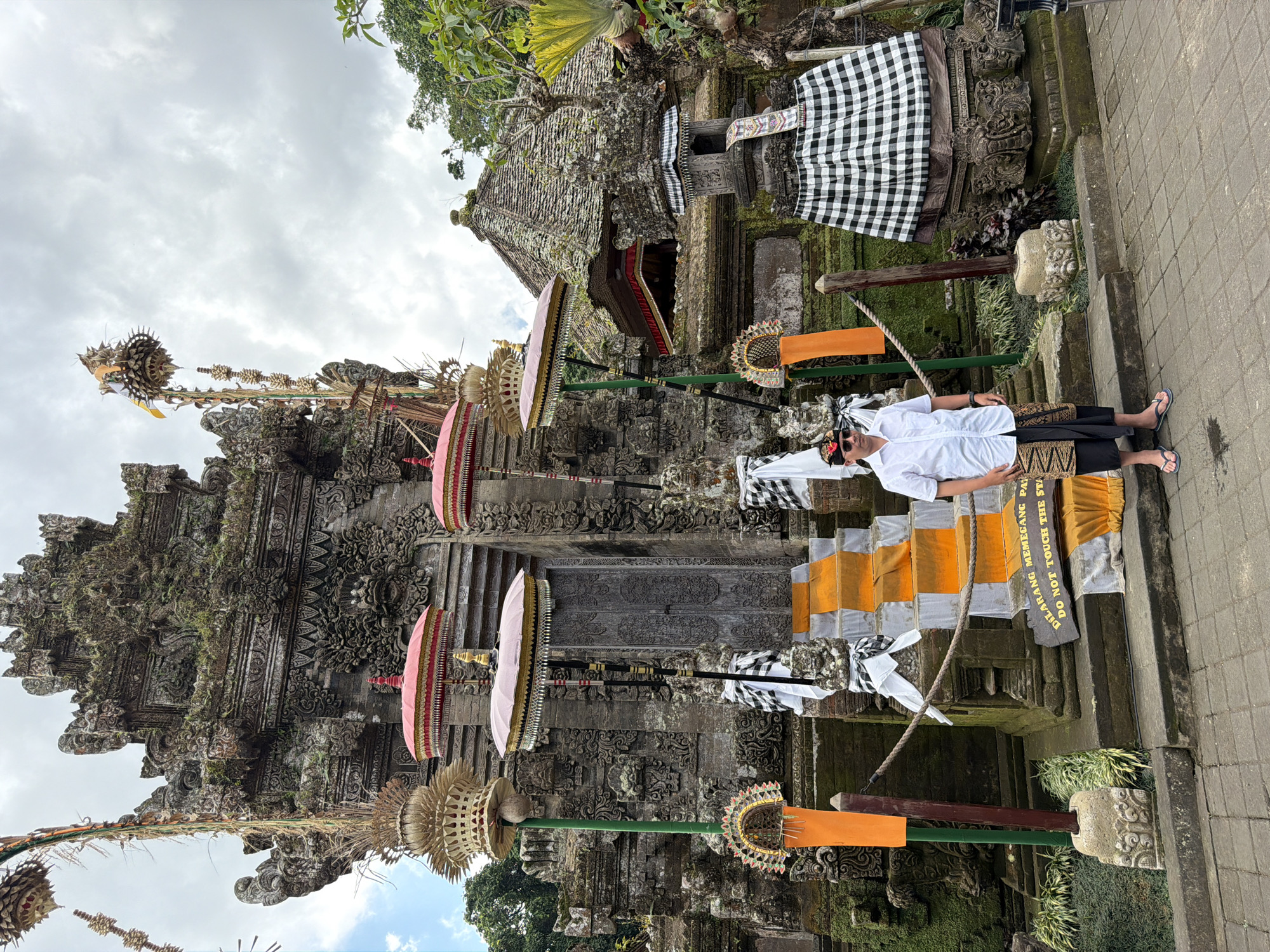 A man in traditional Balinese attire, including a white collared shirt, a sarong, and a head covering, stands in front of a stone temple in Susut, Indonesia. The temple is ornate with intricate carvings and is adorned with decorative elements such as umbrellas, banners, and fabric wrappings. The man is standing on a paved pathway, with steps leading up to the temple entrance. The background features lush greenery and a cloudy sky. The overall scene depicts a cultural and religious setting.