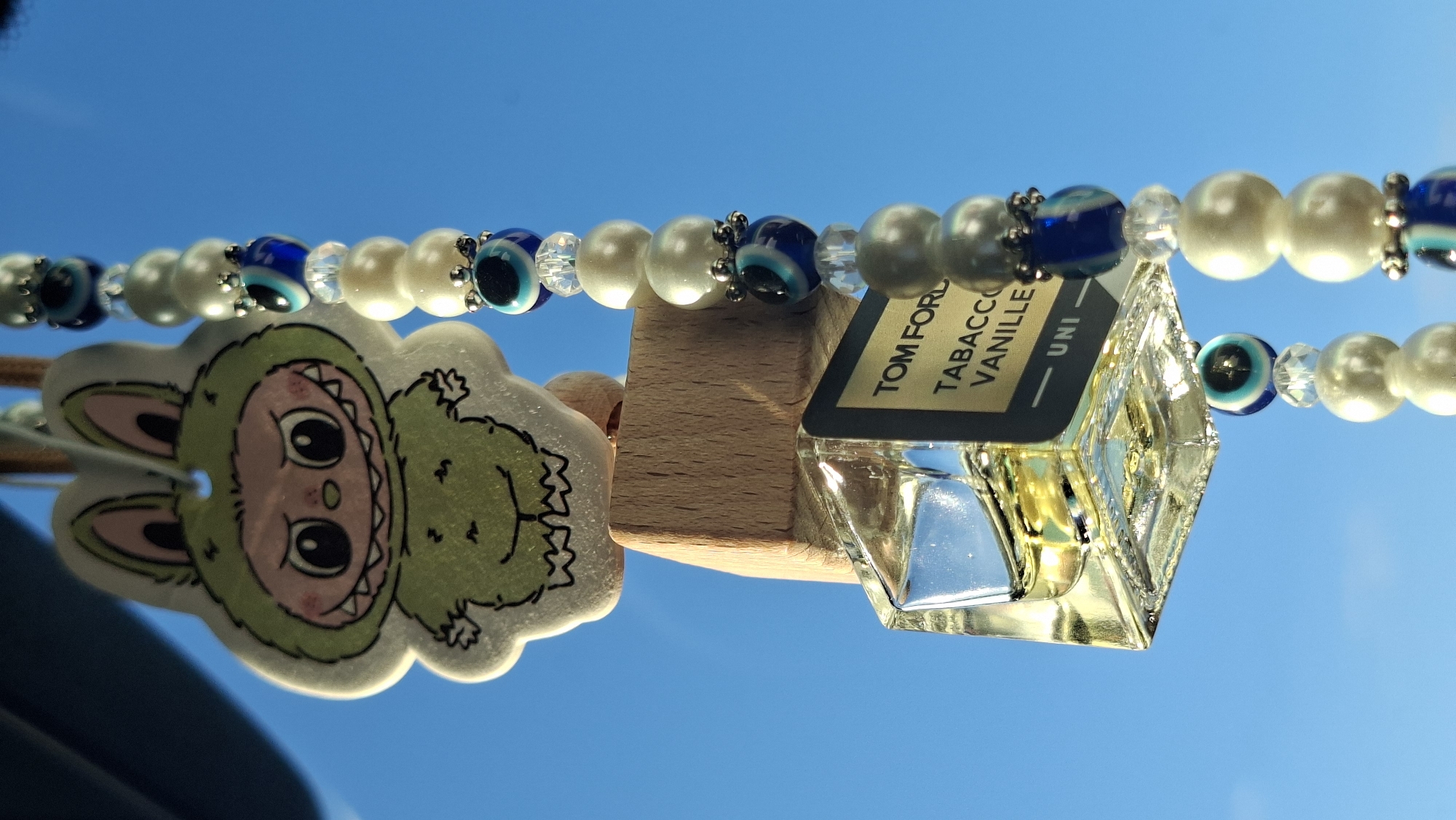 This image shows a close-up of a car air freshener hanging from a rearview mirror. The air freshener is a miniature bottle of Tom Ford Tobacco Vanille perfume. It is hanging from a wooden block that is attached to a string. The string is decorated with white beads and blue and white evil eye charms. Above the perfume bottle, there is a cartoon character of a green monster wearing a bunny costume. The background is a bright blue sky, suggesting it is daytime. The image is taken from a low angle, looking up at the air freshener. There is no discernible location in the city of Smalyavichy, Belarus.