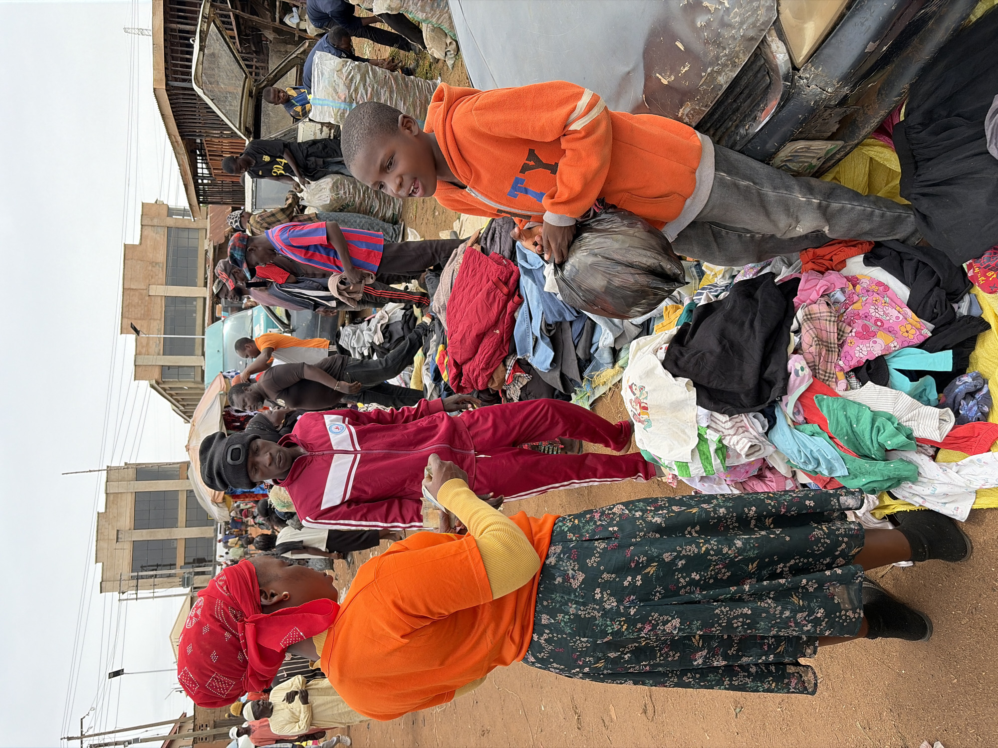This street scene in Gura Top, Nigeria, shows a bustling outdoor market where used clothing is being sold. Several people are present, including adults and children, who are either sorting through piles of clothes or loading them into vehicles.
A young boy wearing an orange hoodie and gray pants is standing on a pile of clothing holding a black plastic bag. Nearby, a woman wearing an orange top, a floral skirt, and a red headscarf is standing on the ground. Another man in a red tracksuit and a black cap is behind her.
Behind the group, other people are seen interacting around piles of clothes. A damaged vehicle is also present, with people loading large white bags or bundles of clothes into it. The buildings in the background suggest an urban environment. The ground appears to be a dusty, dirt surface.