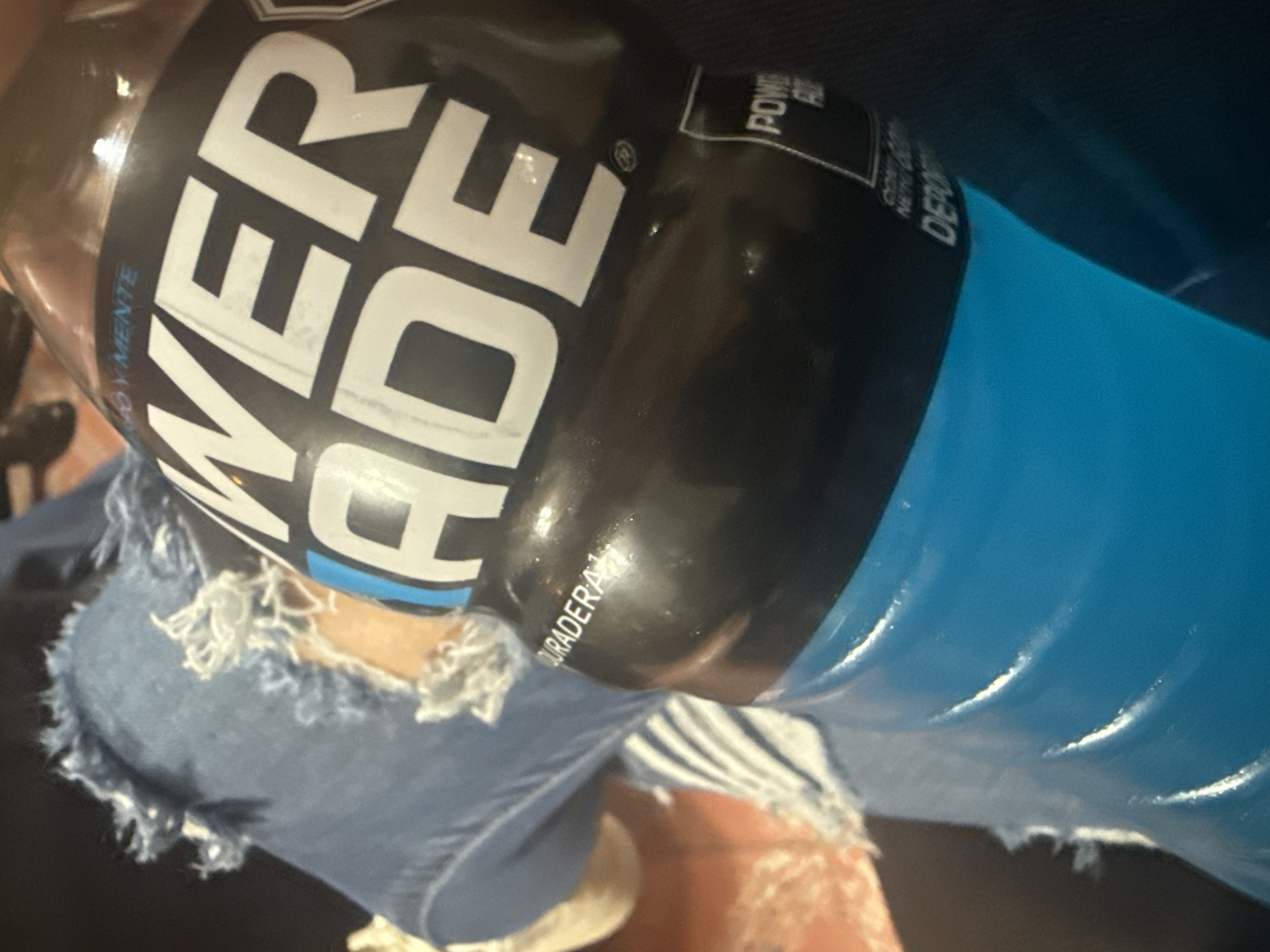 A Powerade bottle with a blue liquid inside rests on a lap covered by distressed denim jeans. The bottle's black label displays the word "POWERADE" in large, white letters. The background is indistinct, but it appears to be indoors. There is no clear evidence to suggest this image was taken in San Cristóbal, Mexico.