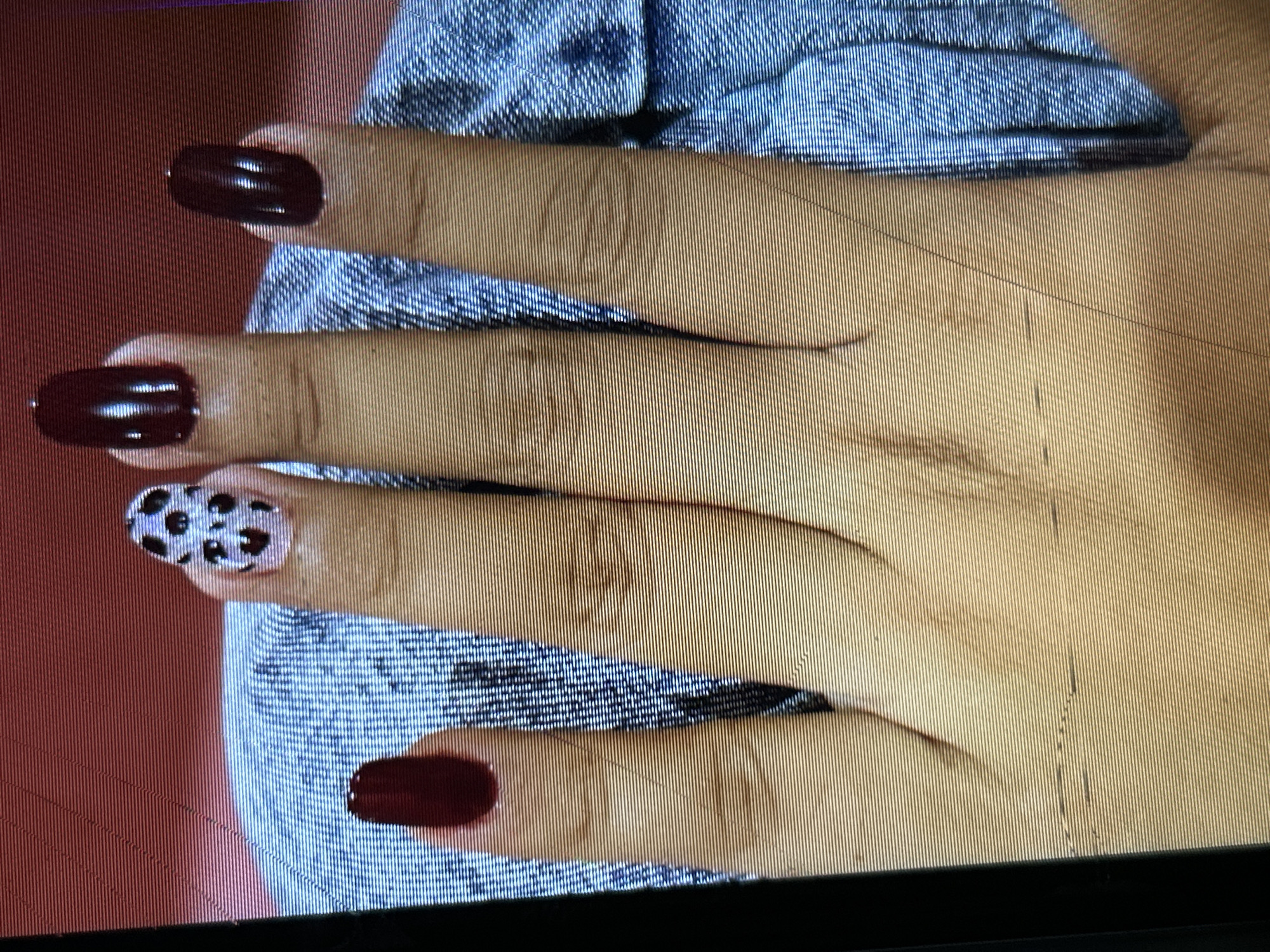 A close-up, slightly blurry image shows a hand with dark red fingernails, except for one nail which has a cow-print design of black spots on a light purple background. The hand is resting on a textured blue fabric that appears to be denim. The background is a solid red, possibly a wall or backdrop. There are no visible elements that can be used to determine the location in Depok, Indonesia. The image is focused on the hand and nails.
