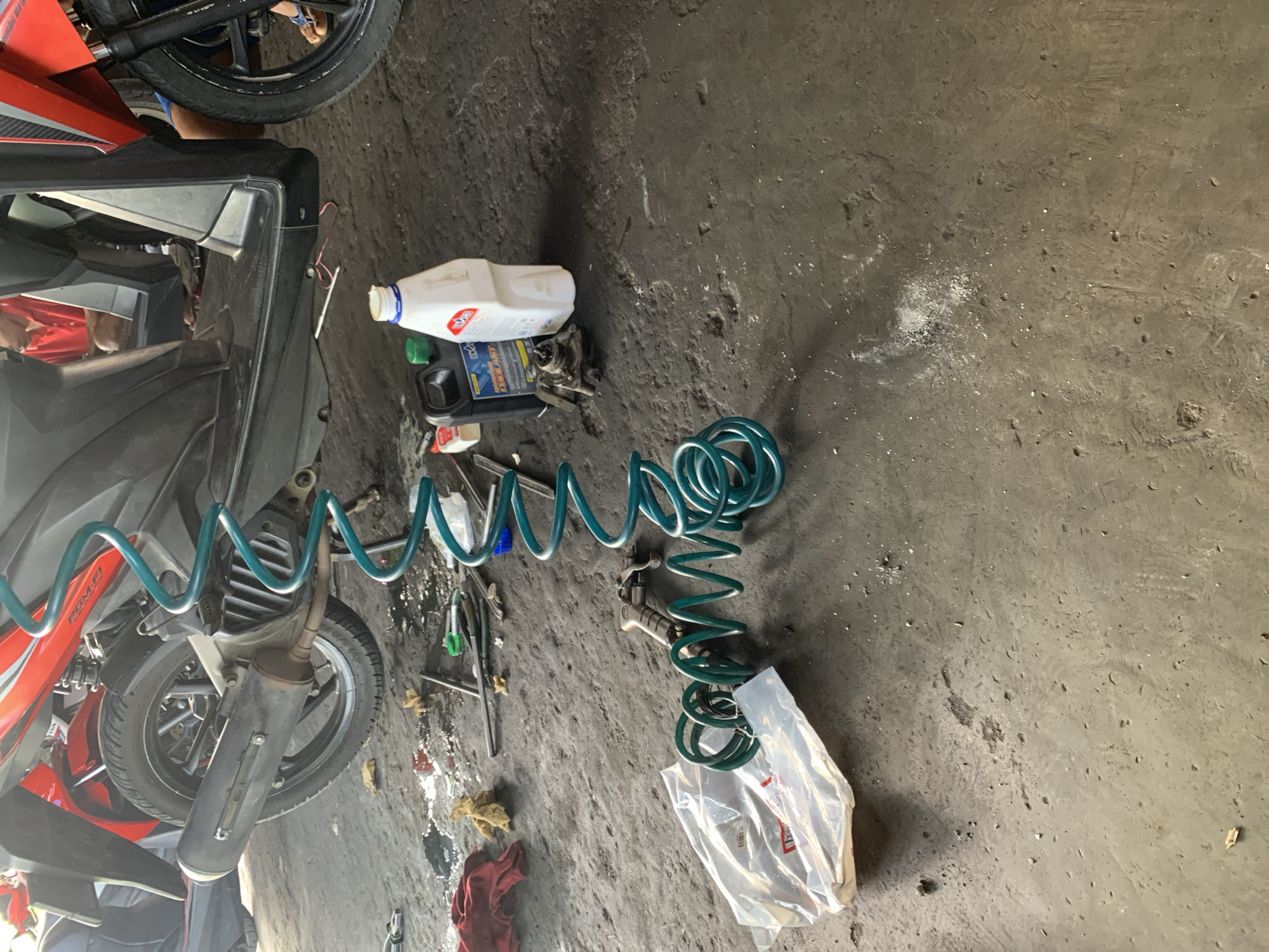 The image depicts a workshop or garage scene in Karangkemiri, Indonesia. A red and black motorcycle is the central subject, partially elevated, revealing its undercarriage. Tools and car maintenance supplies are scattered on the rough concrete floor, which is stained with oil and dirt. Among the items visible are a coiled green air hose, a spray gun, bottles of fluid, and plastic packaging. A person wearing a red shirt is partially visible in the background, suggesting ongoing work or activity within the space. The environment appears to be a utilitarian workspace.