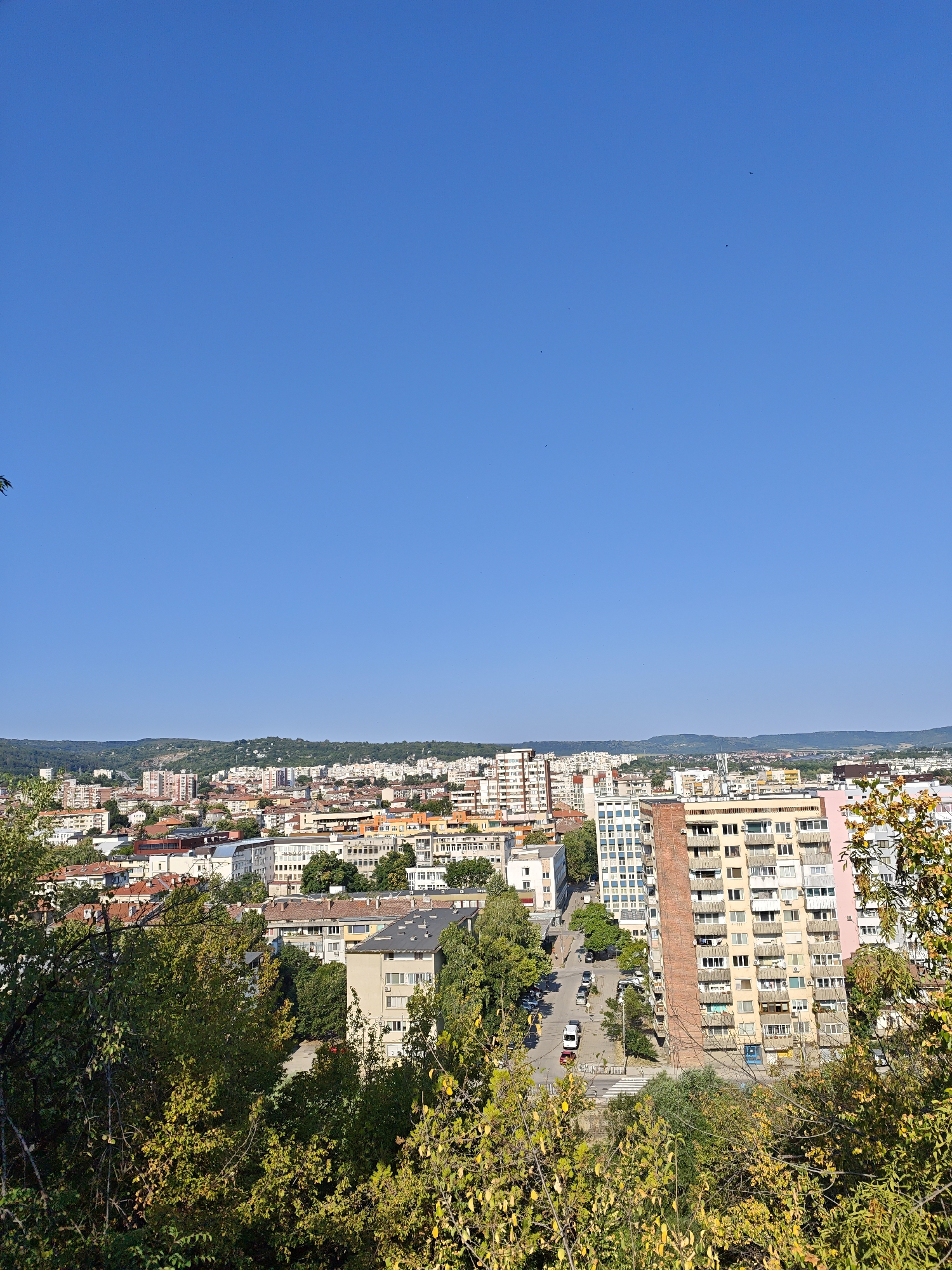 This is an elevated, wide-angle shot of a city with a clear blue sky. The foreground is dominated by lush green and yellow foliage, suggesting an autumn season. Beyond the trees, a dense urban landscape unfolds with a variety of buildings, ranging from smaller, terracotta-roofed structures to taller, multi-story apartment blocks. A street with several parked and moving cars runs through the middle of the scene. In the distance, rolling hills covered in greenery form a backdrop to the city. This view is likely from the Varosha neighborhood in Lovech, Bulgaria, offering a panoramic perspective of the town and its surrounding natural environment.