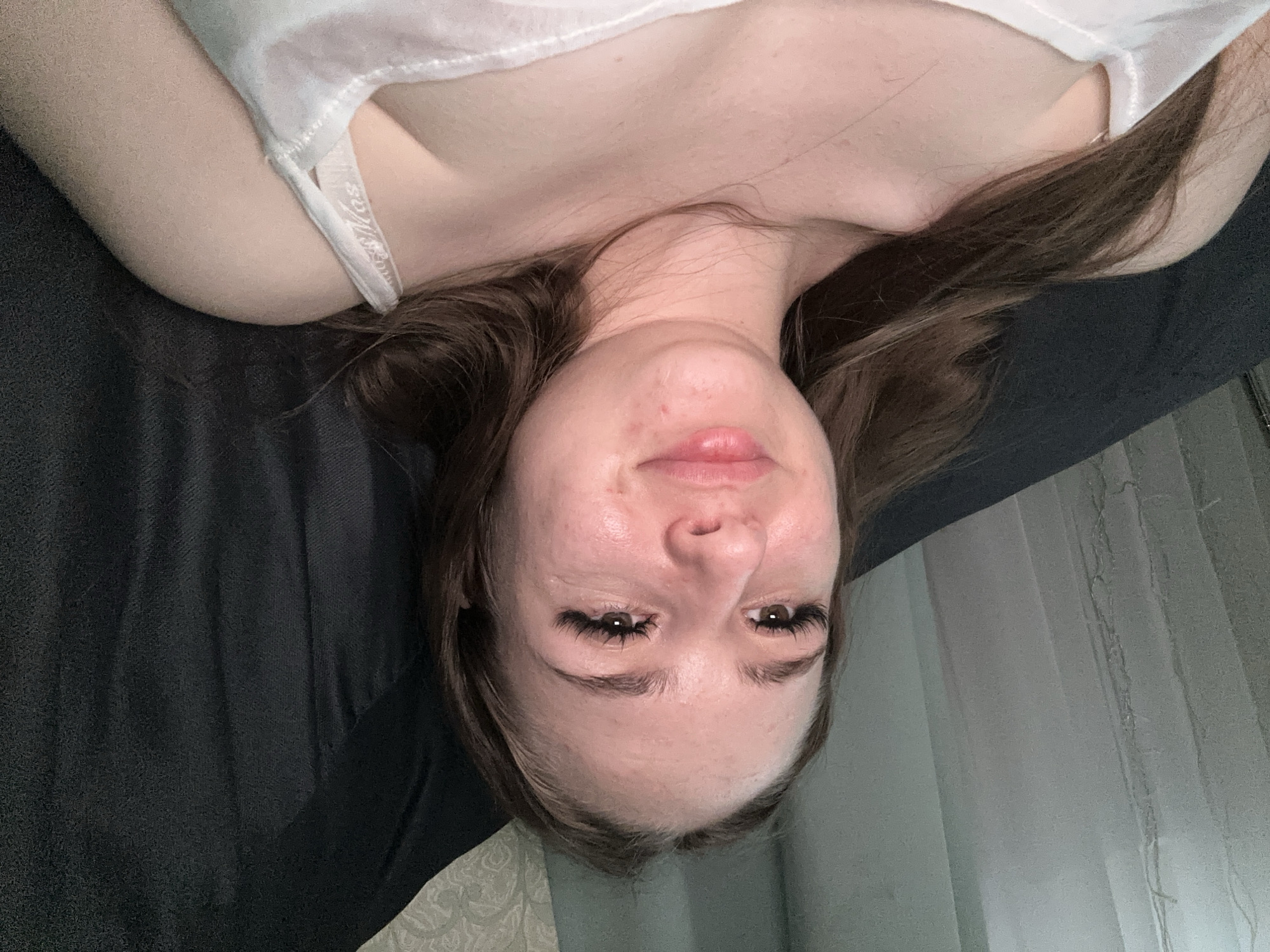 A medium shot captured from a slightly elevated angle shows a young woman with long, brown hair, resting on a dark-colored surface, possibly a couch or bed. She is wearing a white tank top, the strap of which is visible on her right shoulder. Her head is tilted slightly upwards and to her left. The background features sheer, light-colored curtains on the left and a patterned wallpaper on the upper right, suggesting an indoor domestic setting. The lighting appears to be soft and diffused, casting a gentle glow on her face, highlighting her features.