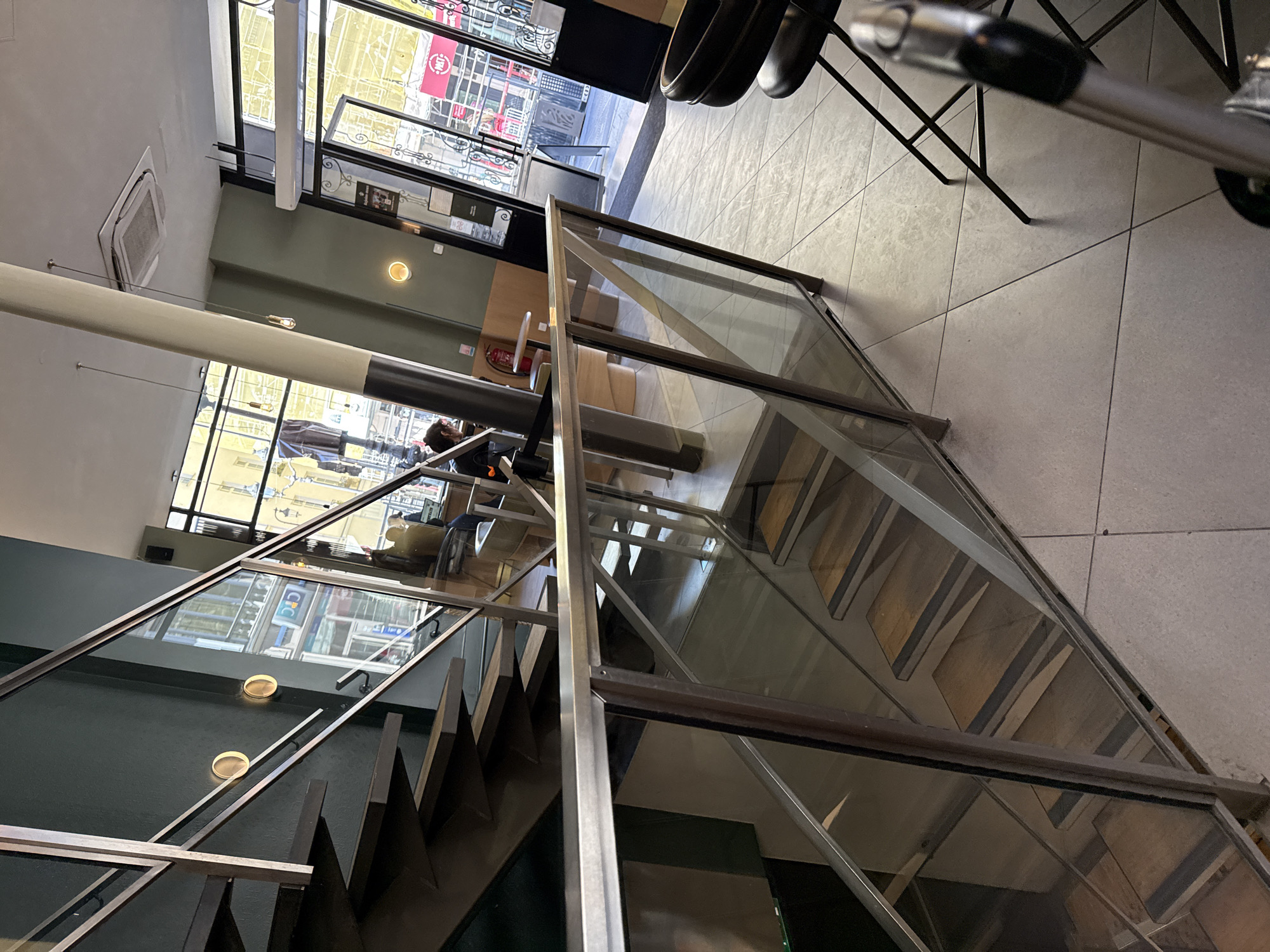 This image depicts the interior of a modern, multi-level establishment, likely a cafe or restaurant, characterized by contemporary design elements. The perspective is taken from an upper level, looking down at a staircase and across a lower floor.
The space is well-lit, suggesting daytime, with ample natural light streaming through large windows that offer a glimpse of an urban street scene outside. The ceiling, visible on the left side of the main image (when viewed upright), features an air conditioning unit and minimalist hanging light fixtures with exposed bulbs. A large white cylindrical pillar extends from the ceiling downwards, and a darker grey pillar is situated further back.
A prominent feature is the staircase, constructed with wooden steps and modern glass railings framed by metal. These railings also extend along an upper walkway or mezzanine. The floor throughout the establishment is covered with light-colored tiles.
On the lower level, seating areas with tables and chairs are visible, including dark-toned chairs and a wooden table. A red fire extinguisher is noticeable near a wall. Reflections in the glass show parts of the exterior, including a red sign with "DM" (or possibly "BM") and another sign displaying "CFC".
A few individuals are present, mostly partially obscured by the structural elements. One person's head is visible near the bottom left, and another is seated near the windows in the background. The overall atmosphere appears calm and inviting.