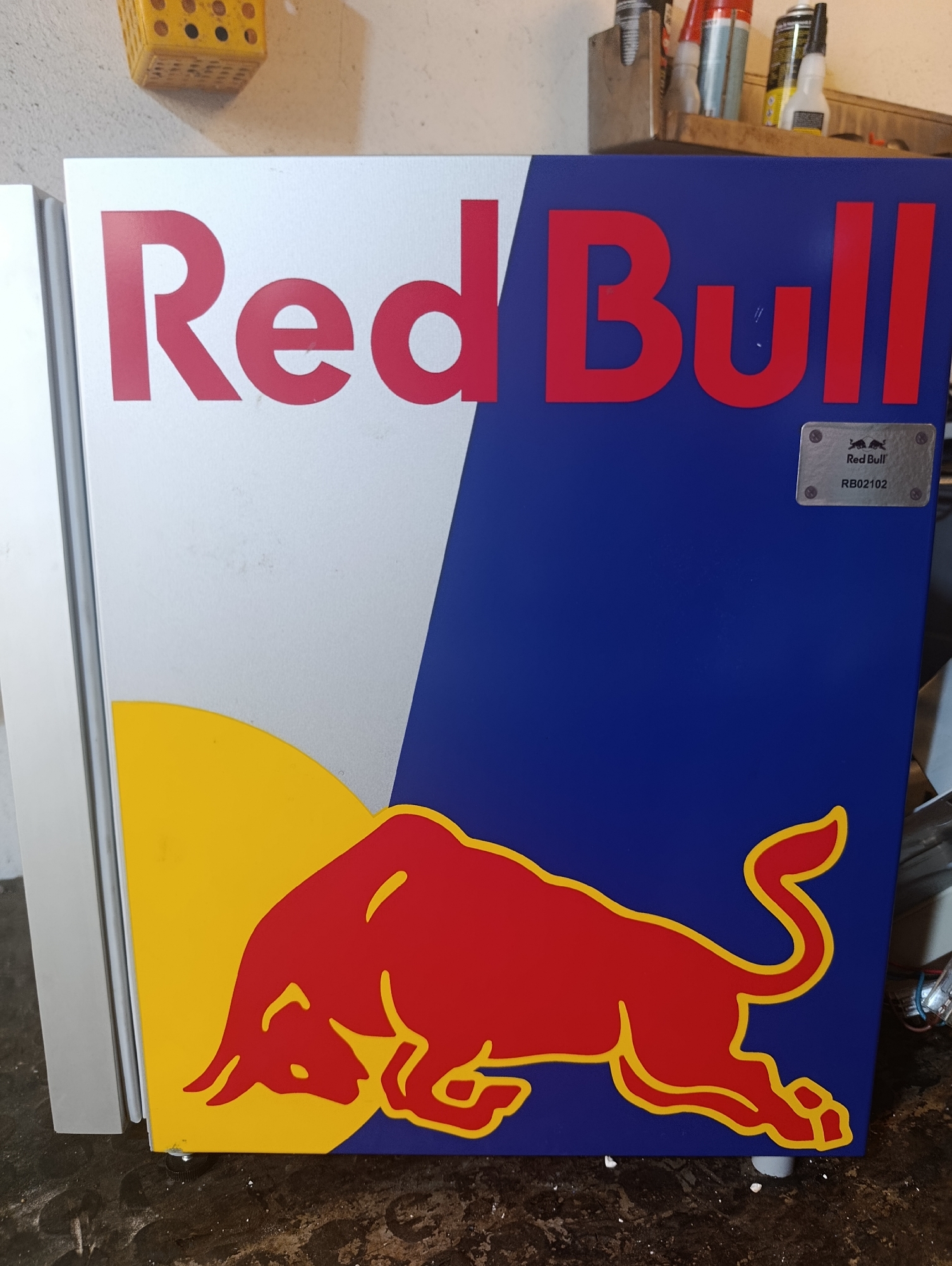 The image shows a close-up of a Red Bull branded mini-fridge. The fridge is predominantly blue with the iconic Red Bull logo featuring two red bulls and the text "Red Bull" in red lettering. A silver plaque with "Red Bull RB02102" is visible on the blue section. The left side of the fridge is silver and features a partial yellow semi-circle and a red bull graphic. The fridge is placed on a dark, textured surface, possibly a floor. In the background, out of focus, there appears to be a shelf with various bottles and containers. The location is identified as Ümraniye, Turkey.