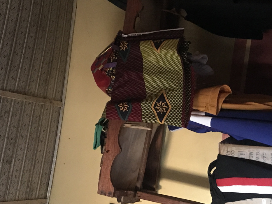 A shelf displays a mix of fabrics and clothing. A colorful, patterned fabric with star-like designs is stacked on top. Below, a glimpse of mustard-colored fabric peeks out from beneath a folded blue and white garment. The shelf's wooden structure has a unique curved design. A glimpse of black fabric with a white and red stripe sits at the bottom. To the left, a patterned metal door or wall is visible.