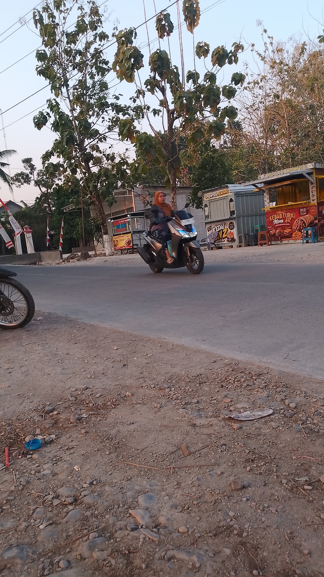 In Karanglangu, Indonesia, a woman wearing a hijab and patterned clothing rides a modern, dark-colored scooter down a paved road. She is moving from left to right in the frame. The road is bordered by a dirt and gravel shoulder on the left, littered with small stones, twigs, and a small blue object. On the right side of the road, a series of food stalls are visible. One stall displays images of food and has the text "KEBAB TURKI MOM AR" on its facade. Another stall to the left has the text "PENTOL KRIWIK" and "KEDAI" visible. Behind the stalls, several trees with sparse foliage are present, and in the background, a clear sky is visible. The overall lighting suggests it is either early morning or late afternoon.