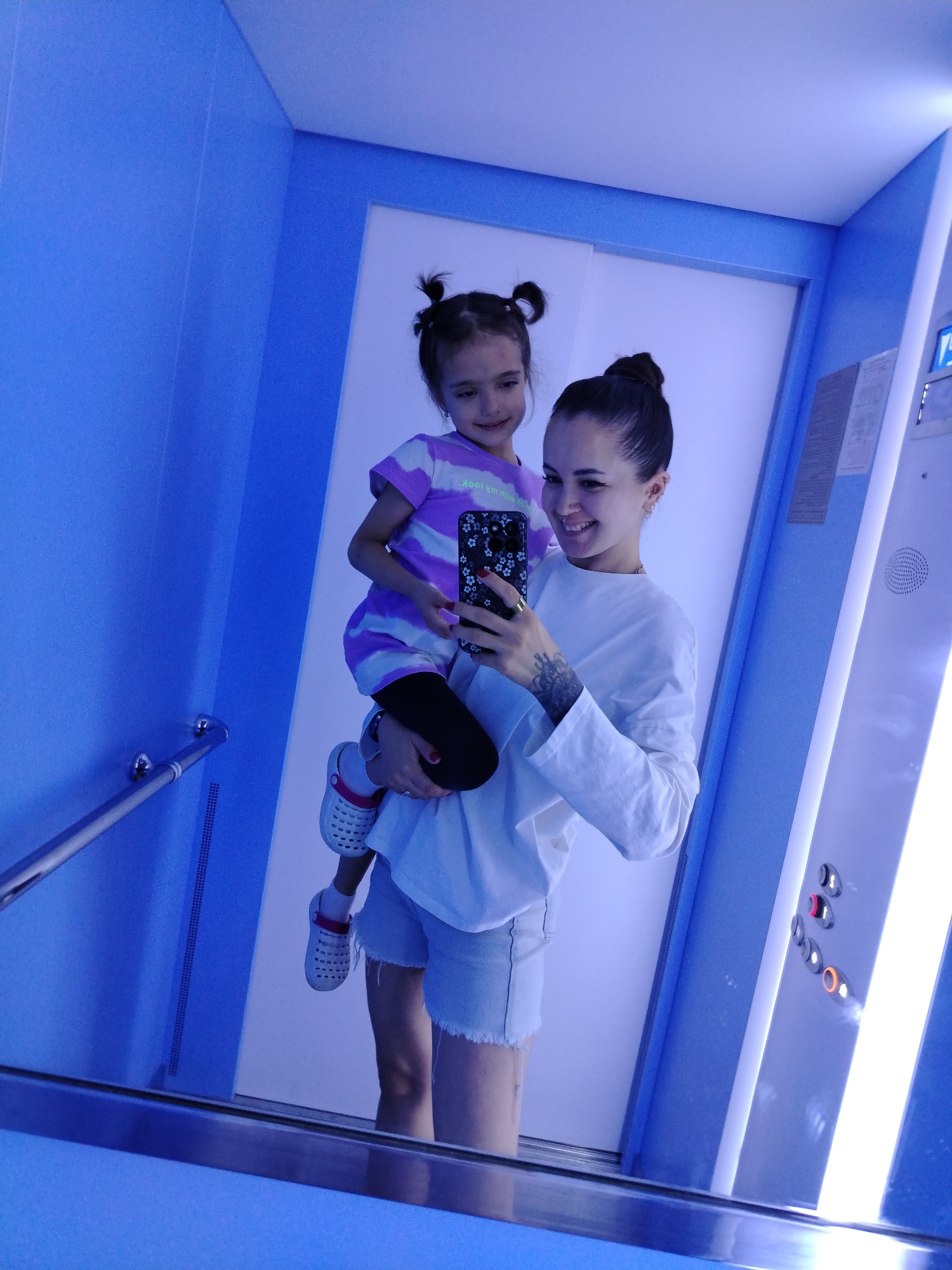 A young woman, a child, and a reflection are visible in what appears to be an elevator. The woman, dressed in a white long-sleeved shirt and denim shorts, is holding the child, who wears a purple and white striped shirt. The woman is taking a selfie with her smartphone. The elevator walls are a vibrant blue, and the floor is reflective, showing a mirror image of the scene. A handrail is mounted on the left wall, and elevator control buttons are visible on the right. The overall lighting is blue-toned. The image was taken in the city of Chaikovsky, Russia.