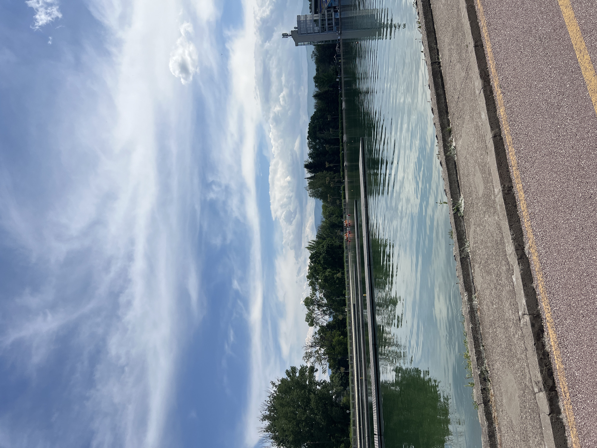 The image shows a view of the Rowing Canal in Plovdiv, Bulgaria. The scene includes the calm water of the canal reflecting the sky, clouds, trees, and a building. Along the edge of the canal is a concrete embankment leading to a paved path with yellow lines. Trees line the opposite side of the canal. The sky is blue with scattered white clouds.