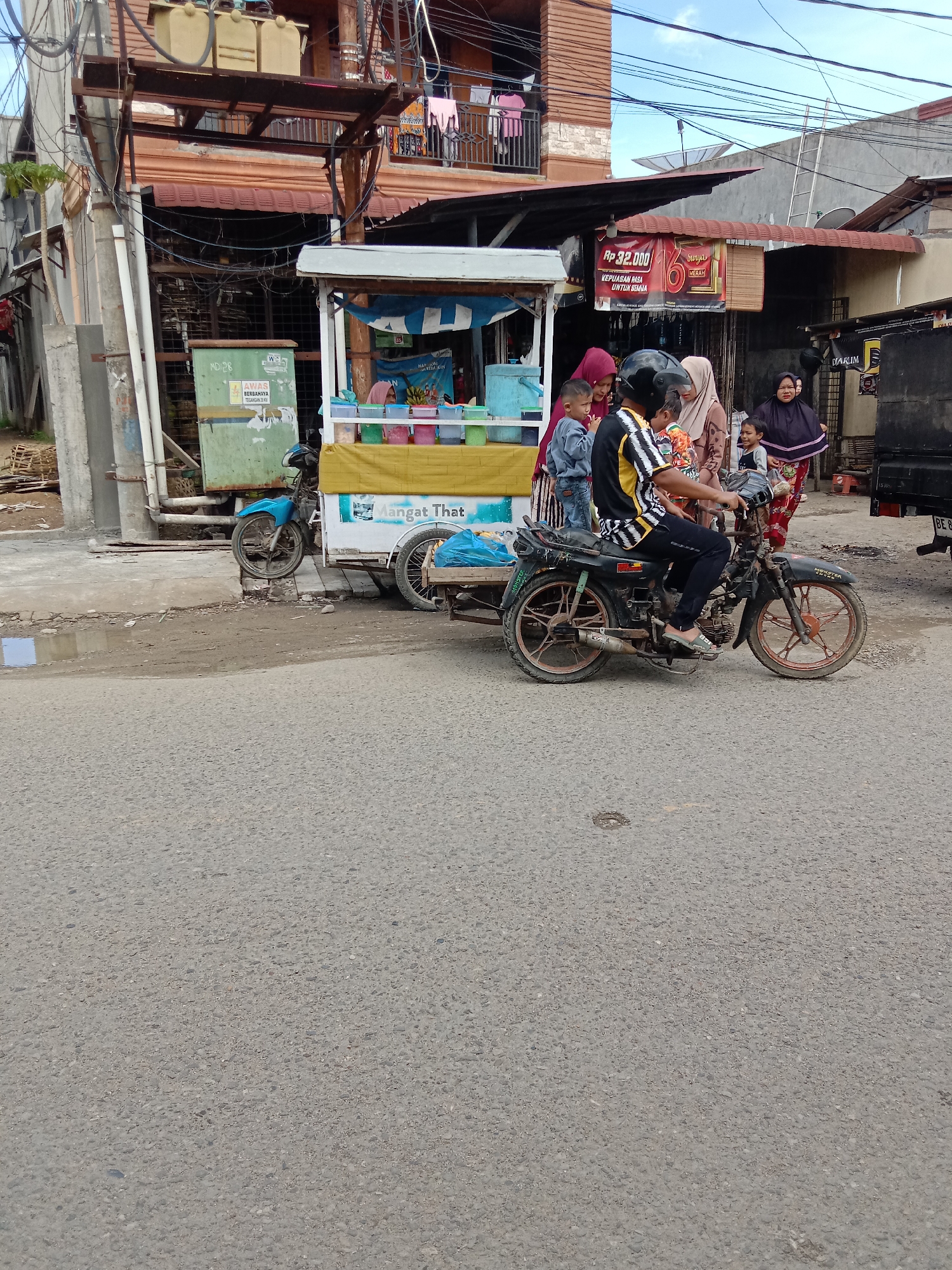 The image captures a street scene in Lhokseumawe, Indonesia. A man wearing a helmet is riding a motorcycle with a sidecar attachment. Several people, including women in headscarves and children, are gathered near a food cart labeled "Mangat That." The cart is parked on the side of the road. Buildings, utility poles, and power lines are visible in the background. There's a poster with the price "Rp 32.000" and the words "Surya Merah." A dark-colored truck is partially visible on the right side of the frame. The road surface is asphalt.