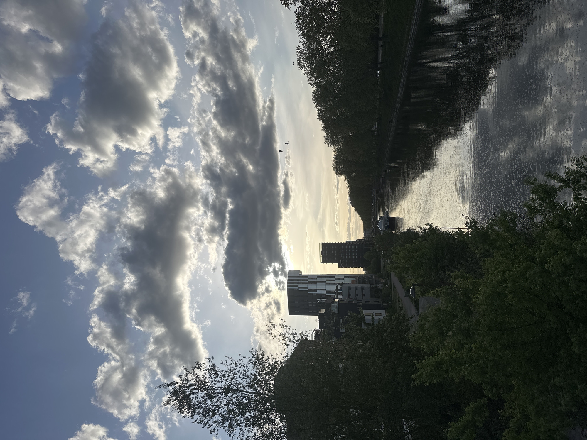 This image captures a view in Strasbourg, France. The scene includes a body of water, possibly a canal or the Ill River, with trees lining its banks on both sides. Modern buildings are visible in the background, partially obscured by foliage. The sky is filled with clouds, casting reflections on the water's surface. The photograph appears to have been taken during the daytime, likely in the late afternoon or early evening, as suggested by the angle and intensity of the light. The composition is such that the sky and clouds occupy a significant portion of the frame, emphasizing the natural elements of the scene. Birds in the sky are barely visible.