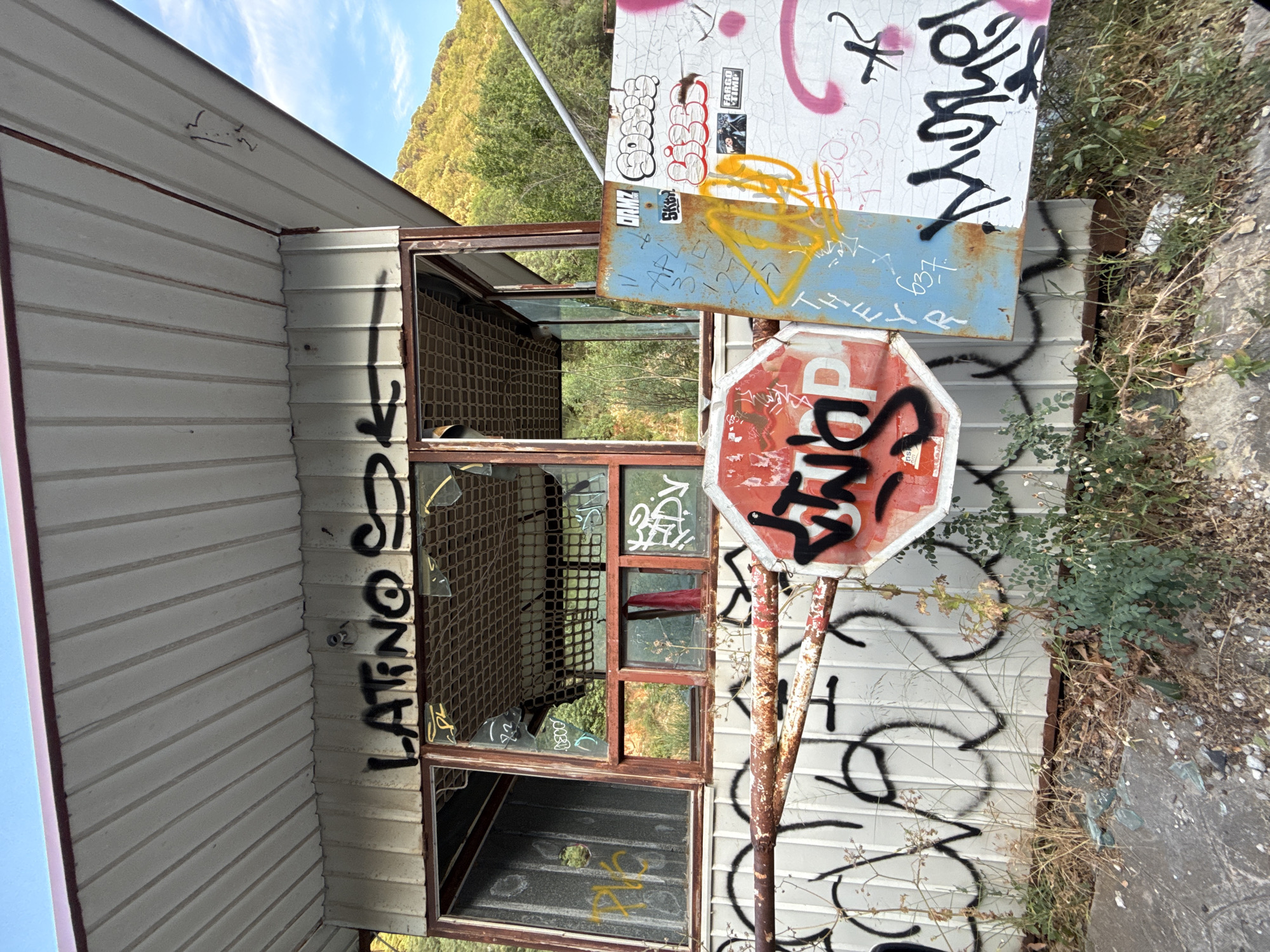 The image depicts an abandoned, graffiti-covered structure in Perazića Do, Montenegro, under a clear blue sky. The structure, possibly a former toll booth or guard house, has a corrugated metal roof and siding. Its windows are broken and boarded with wire mesh. Numerous spray-painted tags and graffiti adorn the walls, a rusted metal gate, and various signs attached to the structure. These signs include a red octagonal stop sign with black graffiti, a blue metal sign with handwritten text and yellow spray paint, and a white board with multiple graffiti tags and stickers, one of which reads "FARGO TIME". Overgrown vegetation and debris are present at the base of the structure, indicating its derelict state. The background features a hillside covered with trees, suggesting a semi-rural or natural setting within the town.