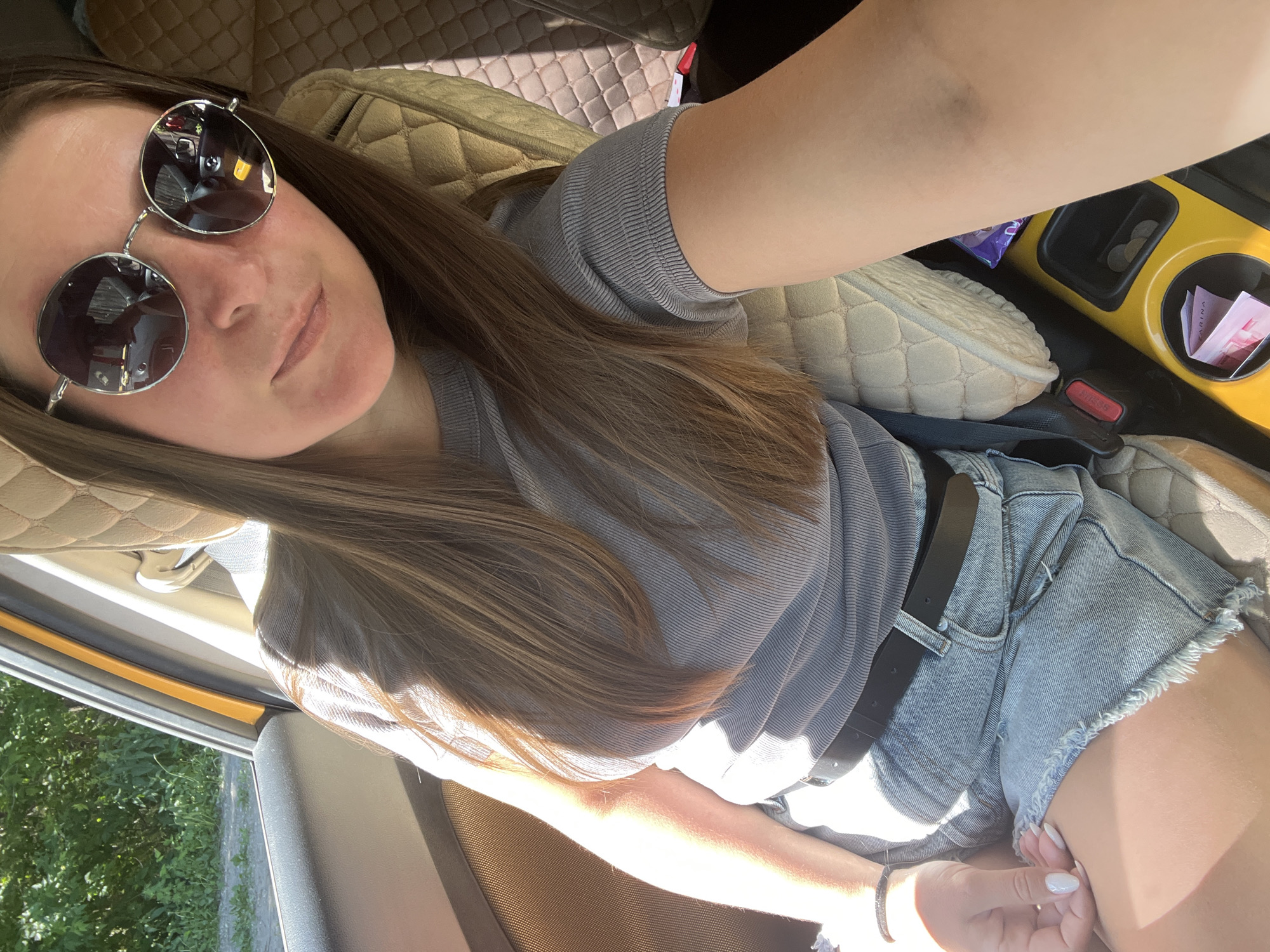 The image shows a young woman sitting in the passenger seat of a car. She is wearing a grey t-shirt, denim shorts, and round sunglasses. The car interior appears to be a light beige color, with quilted upholstery on the headrest and seat. Sunlight streams into the car, casting shadows and highlighting the woman's hair and arm. The background outside the car window shows greenery, suggesting a natural or suburban environment, possibly on the outskirts of Novosibirsk, Russia. The woman is looking at the camera, and her arm is resting on her leg. A black seatbelt is visible, as is a cup holder with some coins and a folded piece of paper inside. The overall impression is a candid snapshot taken during a car journey.