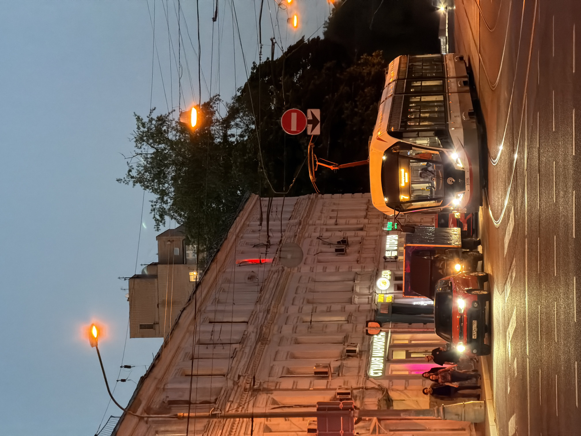 The image captures a street scene in Moscow, Russia, during twilight. A modern tram with illuminated front displays is prominently featured, traveling along a wet, reflective road. To the right of the tram, two parked cars are visible. Along the left of the frame is a light-colored building with multiple stories and illuminated signs indicating businesses. Beneath the signs, a group of people are standing or sitting on a low wall. Streetlights are spaced along the road, emitting an orange glow. Overhead, a network of wires intersects the sky. Road signs are also visible. The background shows a tree line and glimpses of other buildings under a dusky sky.