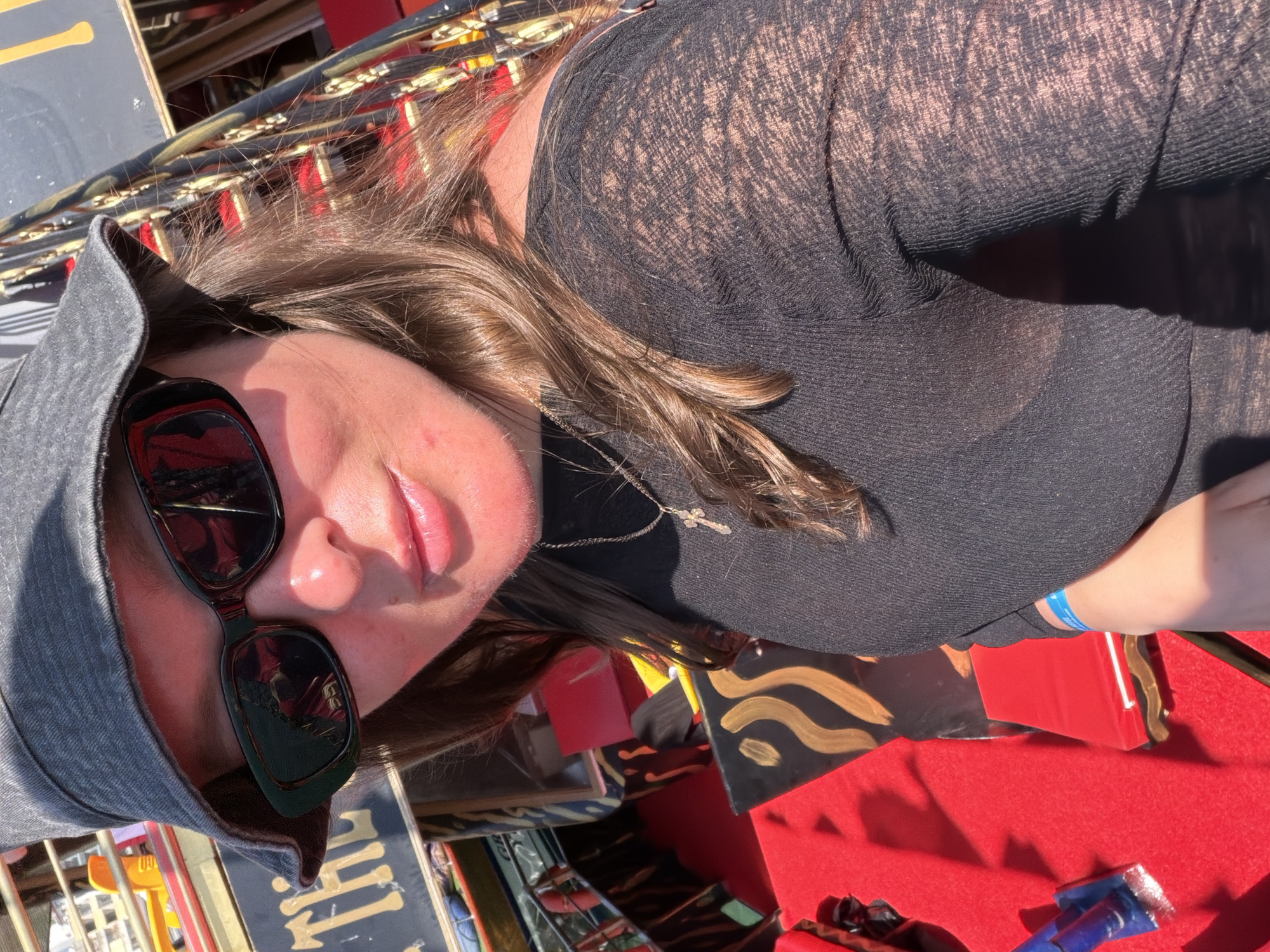 The image shows a person wearing sunglasses and a gray hat, with a black top that reveals some skin. A silver necklace with a cross pendant is visible. The person's arm has a blue wristband. The foreground includes a red surface, and behind that are various objects with patterns and colors, possibly merchandise on display. The location appears to be outdoors, possibly a market or fair in Alanya, Turkey, due to the items for sale and the general setting.