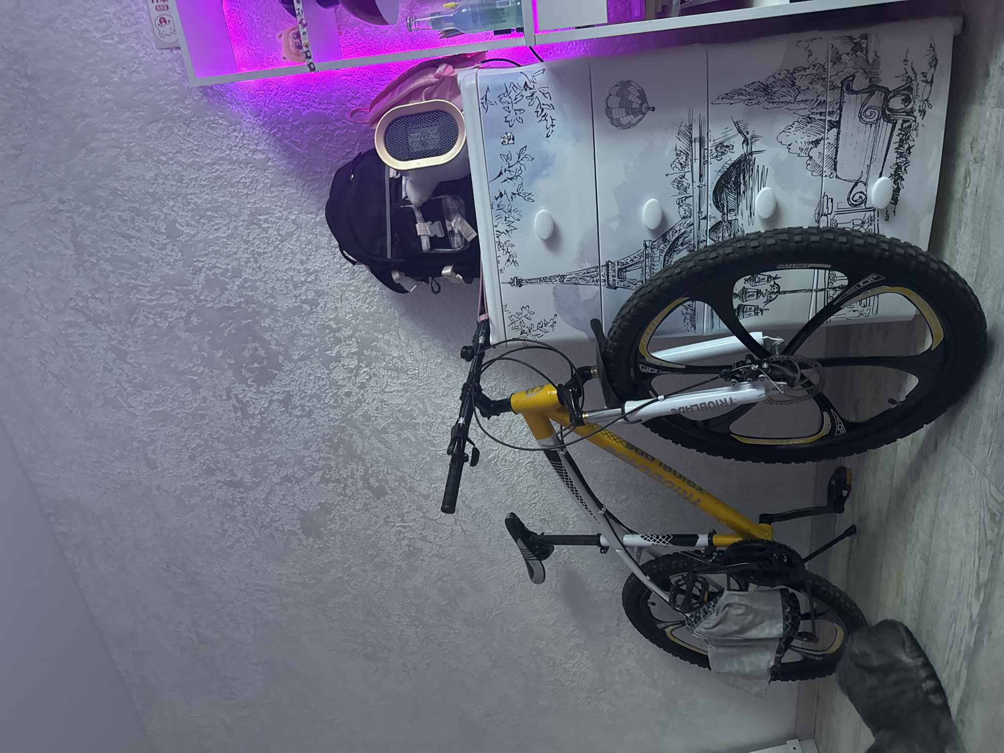 The image shows a yellow and white mountain bike leaning against a wall. The wall has a textured finish. To the right of the bike is a white dresser decorated with a black and white sketch of Parisian landmarks, including the Eiffel Tower and hot air balloons. On top of the dresser are a black backpack and a small, oval-shaped heater. A pink backpack is also visible. A shelf unit to the far right has purple LED lighting and several items, including what appear to be bottles and small decorative objects. A grey cat is visible in the lower left corner of the image, facing away from the camera. The floor is a light-colored material.